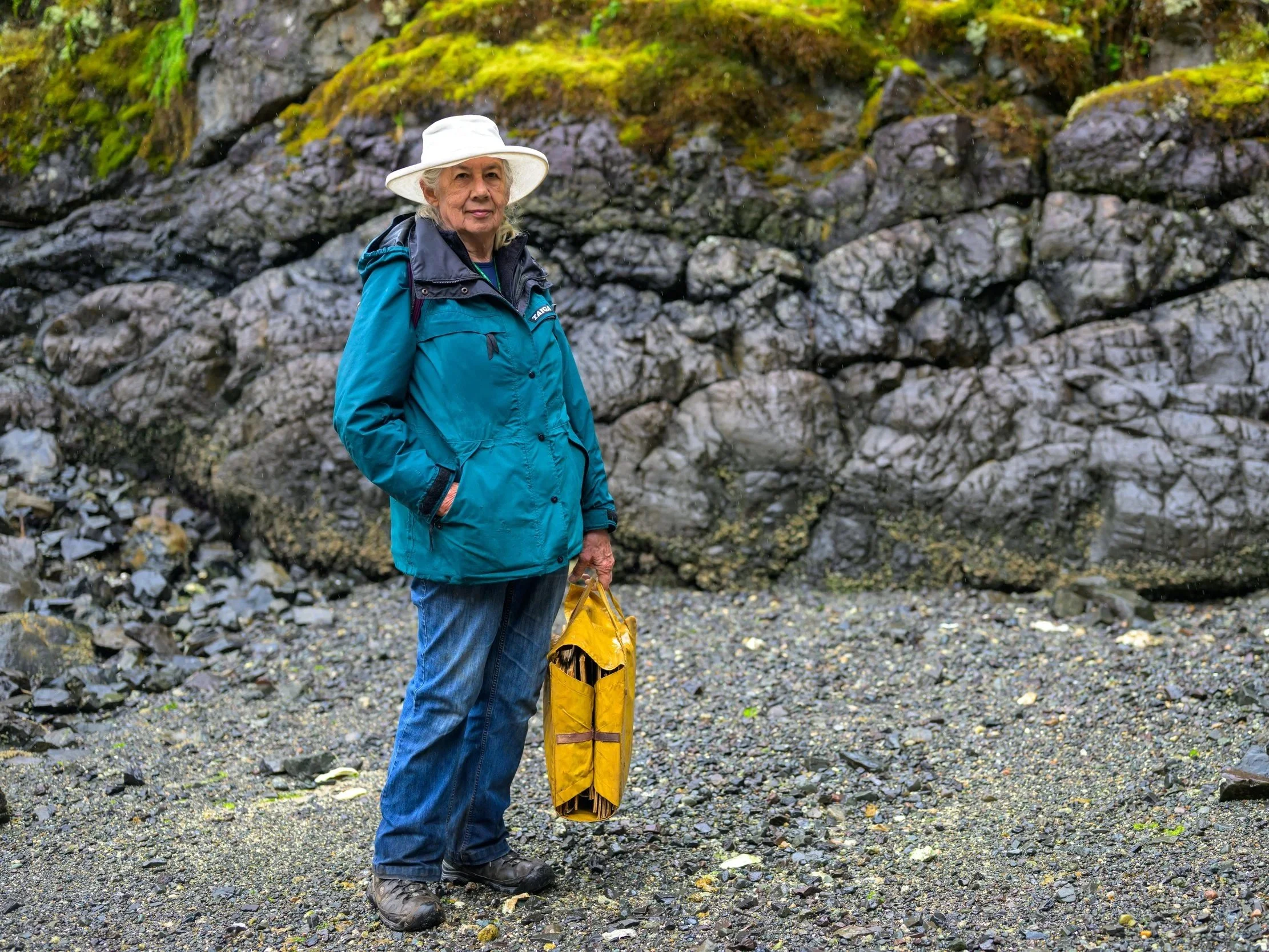An elderly woman standing on a rocky, pebbly beach wearing a teal jacket, jeans, hiking boots, and a white hat, holding a yellow bag, with a moss-covered rock formation in the background.