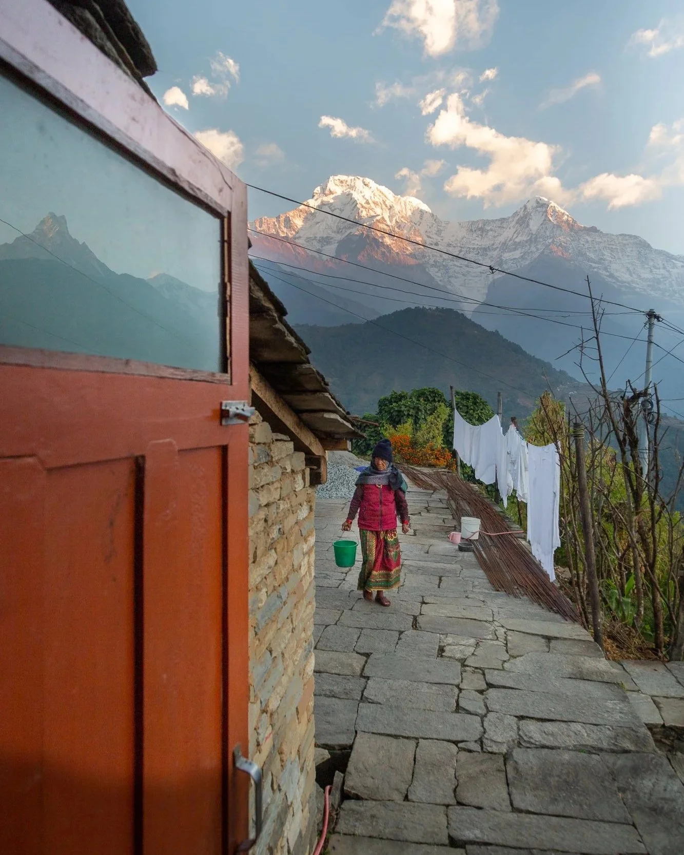A woman walking on a stone pathway in a mountain village with snow-capped peaks in the background, laundry hanging on a line, and a small building with a wooden door in the foreground.