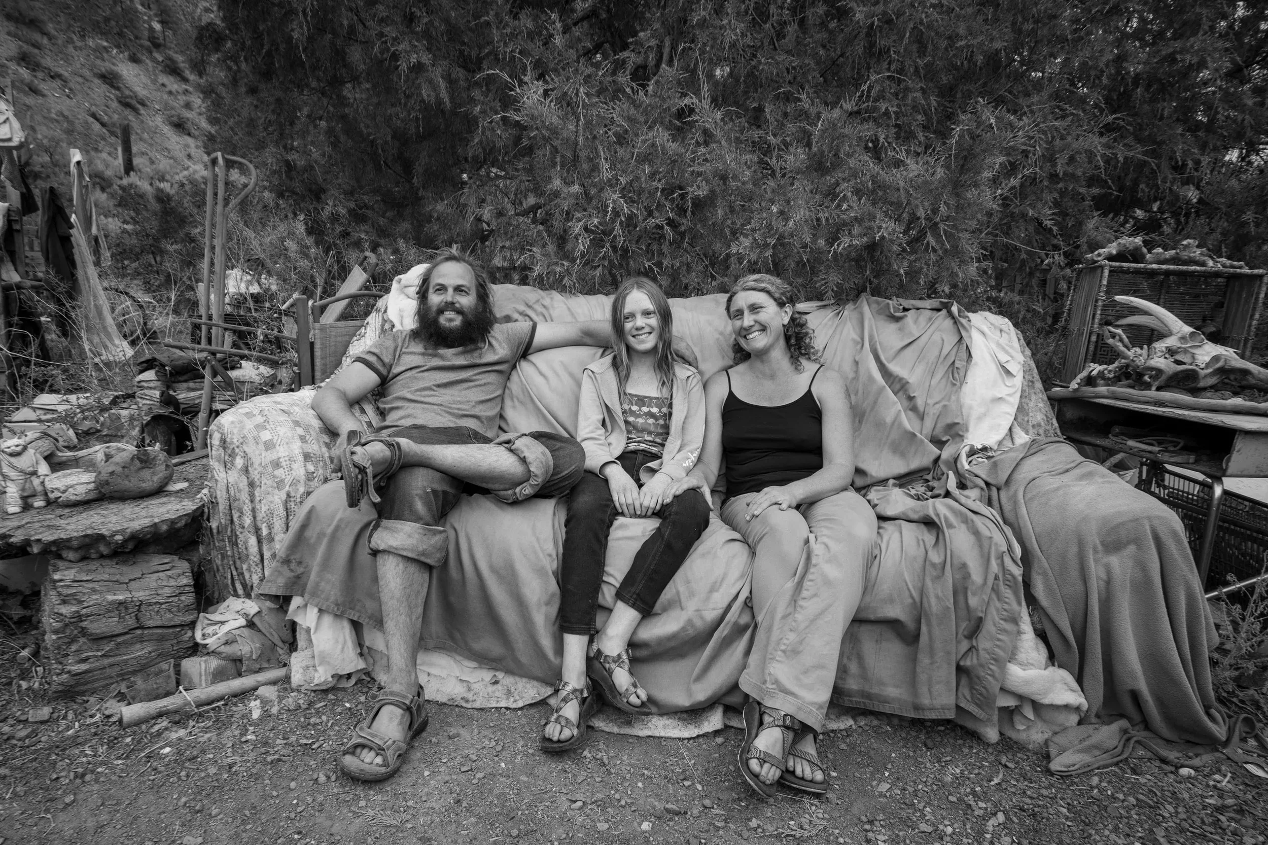 Three people sitting on a makeshift sofa outdoors, smiling and looking at the camera. The background contains trees and various objects, including animal skulls and bones.