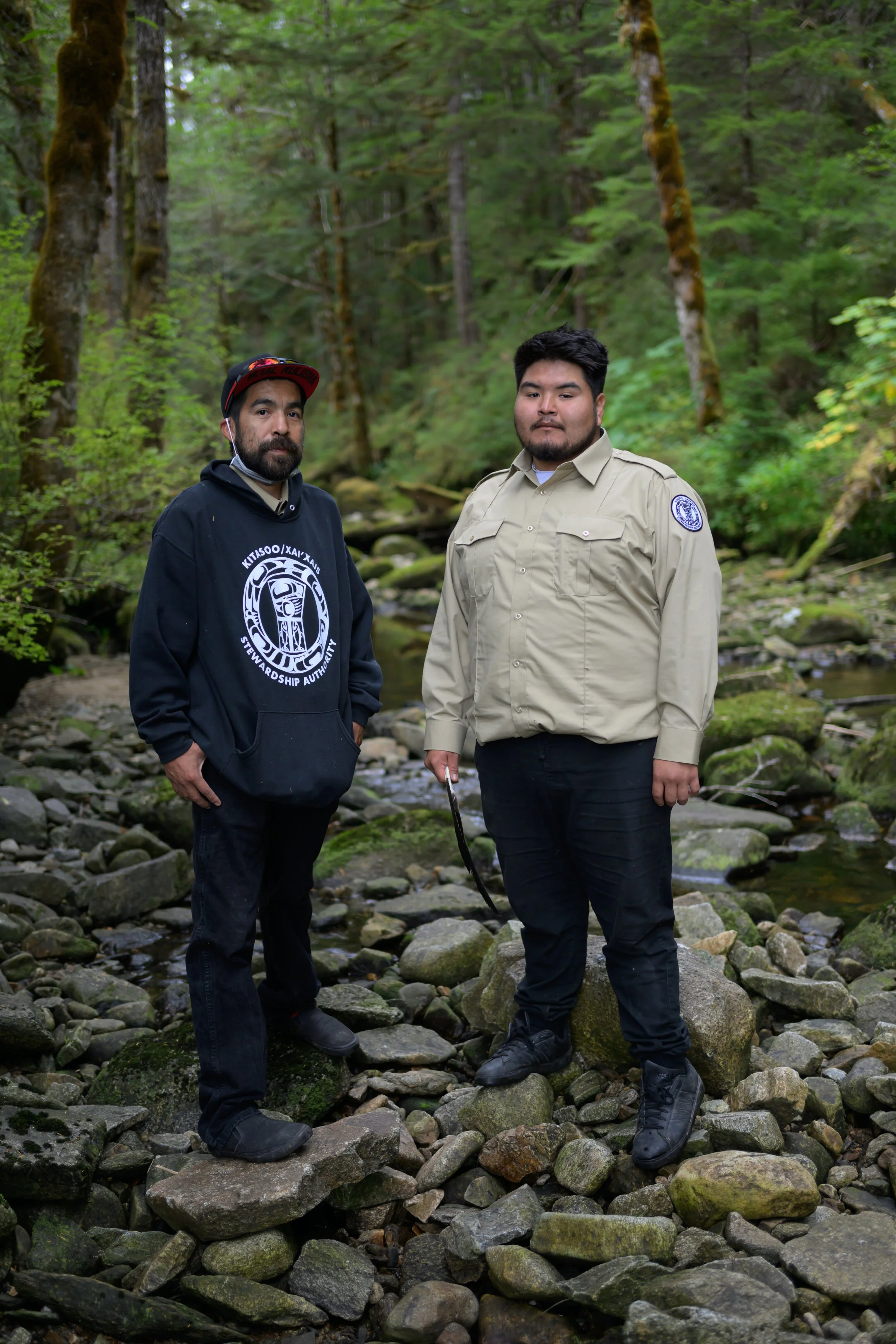 Two men standing on rocky terrain in a forest, with trees and a small stream in the background. One is wearing a black hoodie with a logo, and the other is in a beige uniform shirt with a patch on the sleeve, holding a stick.