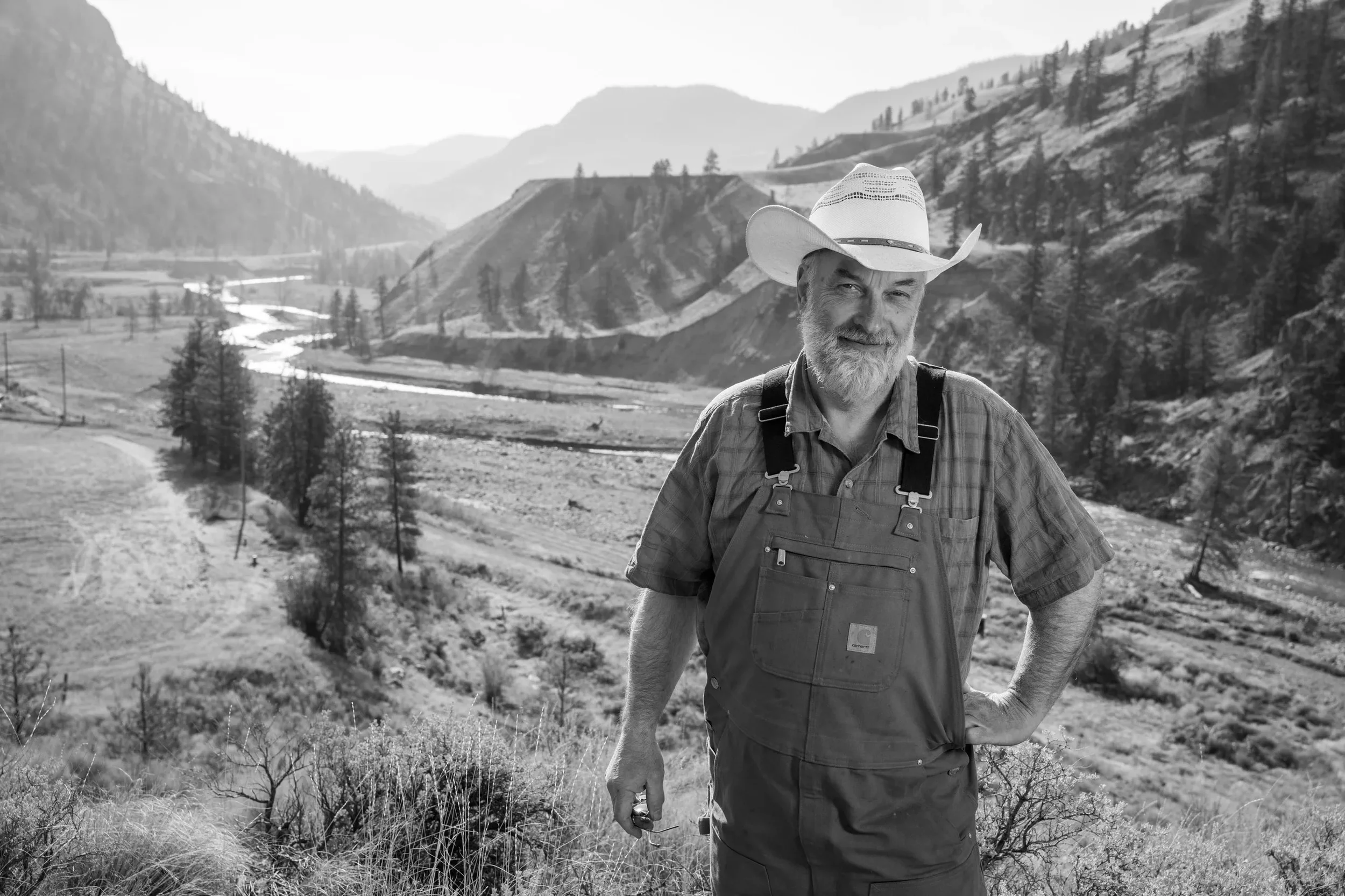 A man with a cowboy hat and beard standing outdoors in a mountainous landscape with valleys, trees, and hills in the background