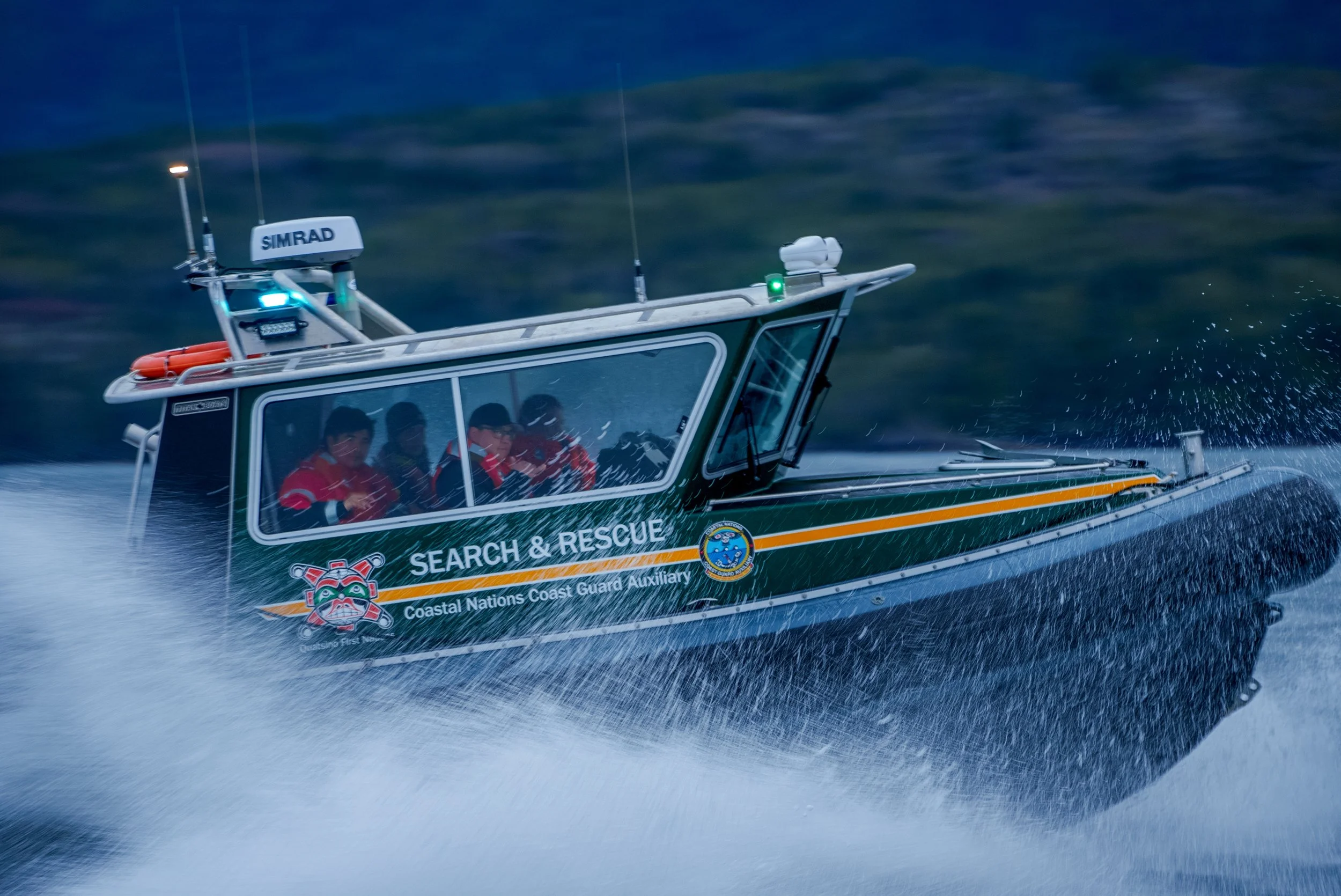 A Coast Guard search and rescue boat speeding through the water, creating spray, with four crew members inside visible through the windows, wearing life jackets and gear.