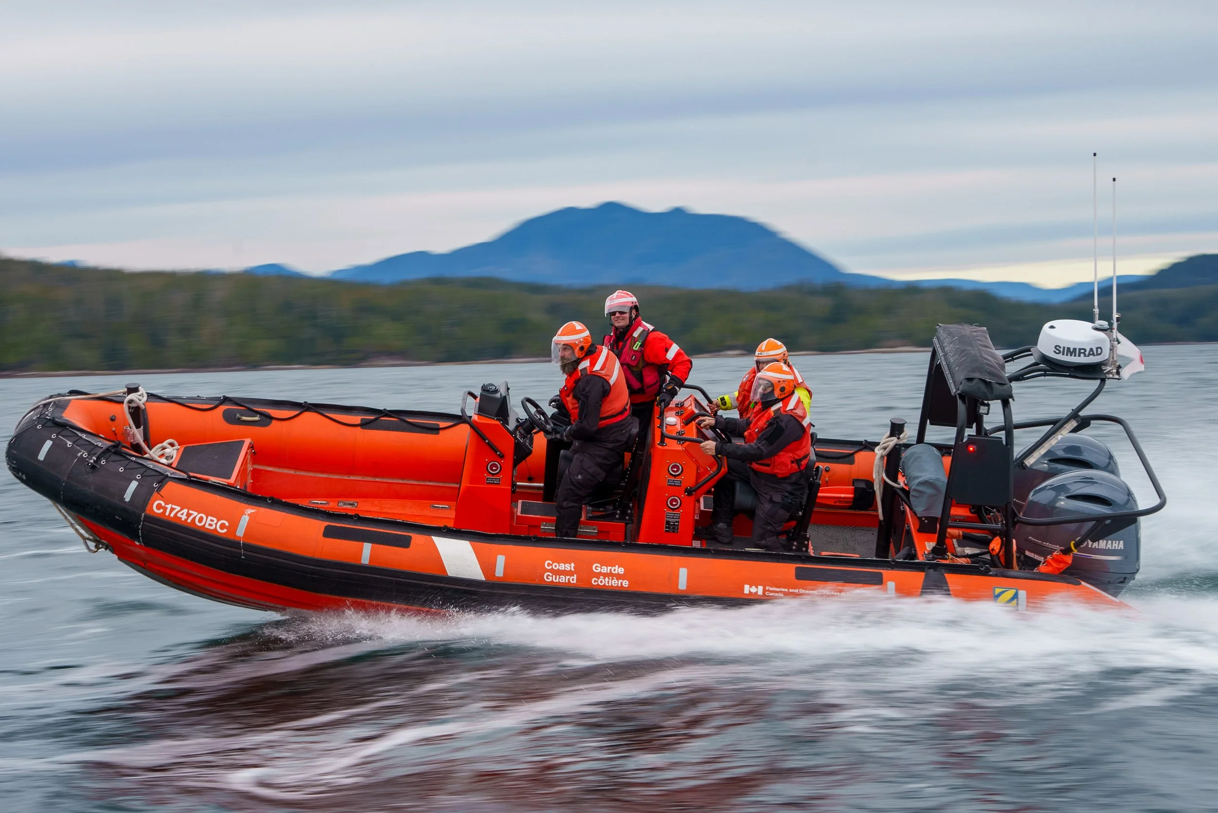 A Canada Coast Guard inflatable boat with four crew members wearing orange helmets and life jackets, operating on a body of water with a mountainous landscape in the background.