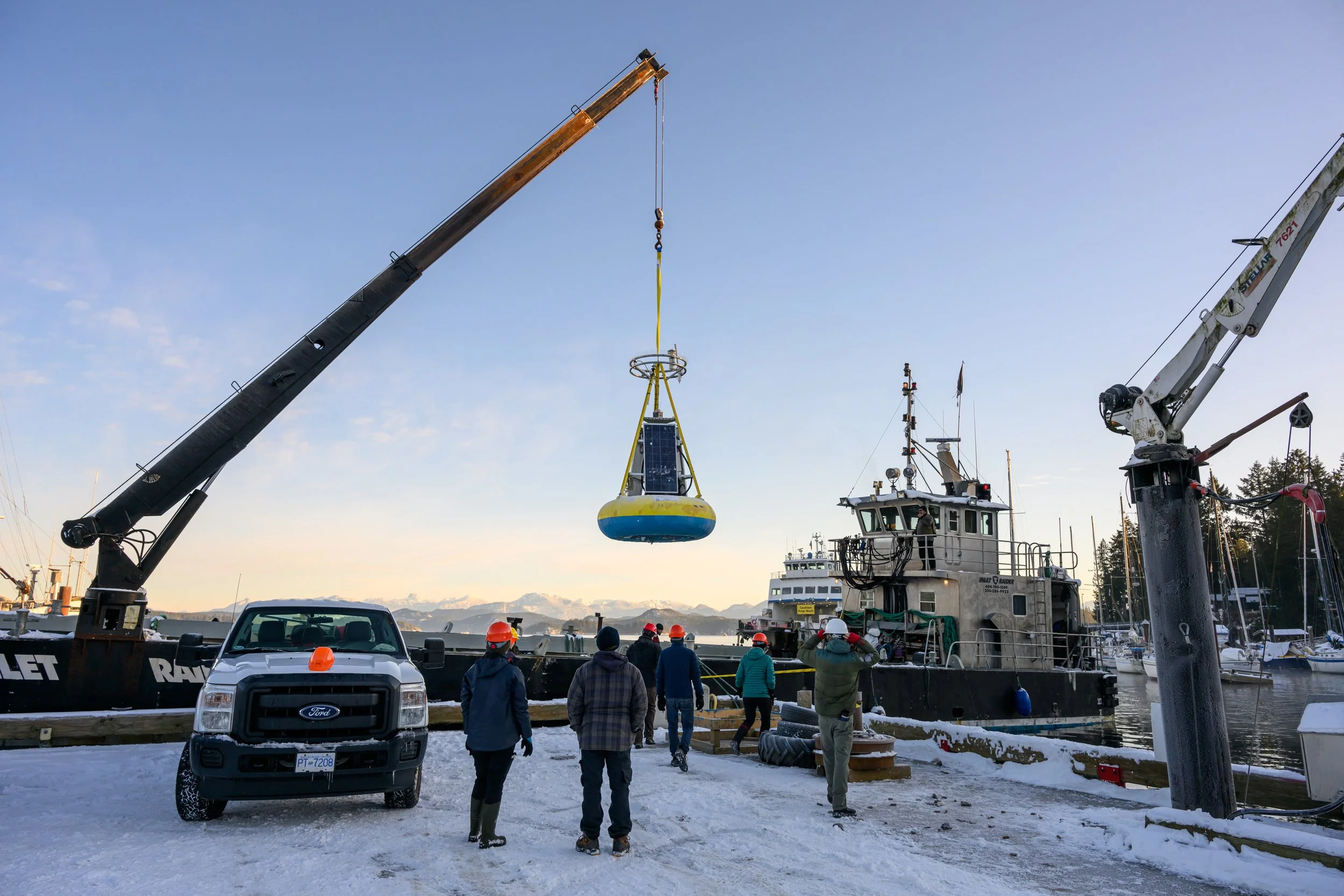 Workers and engineers on a snowy dock observing a large buoy being lifted by a crane from a boat, with sailboats and snow-capped mountains in the background.