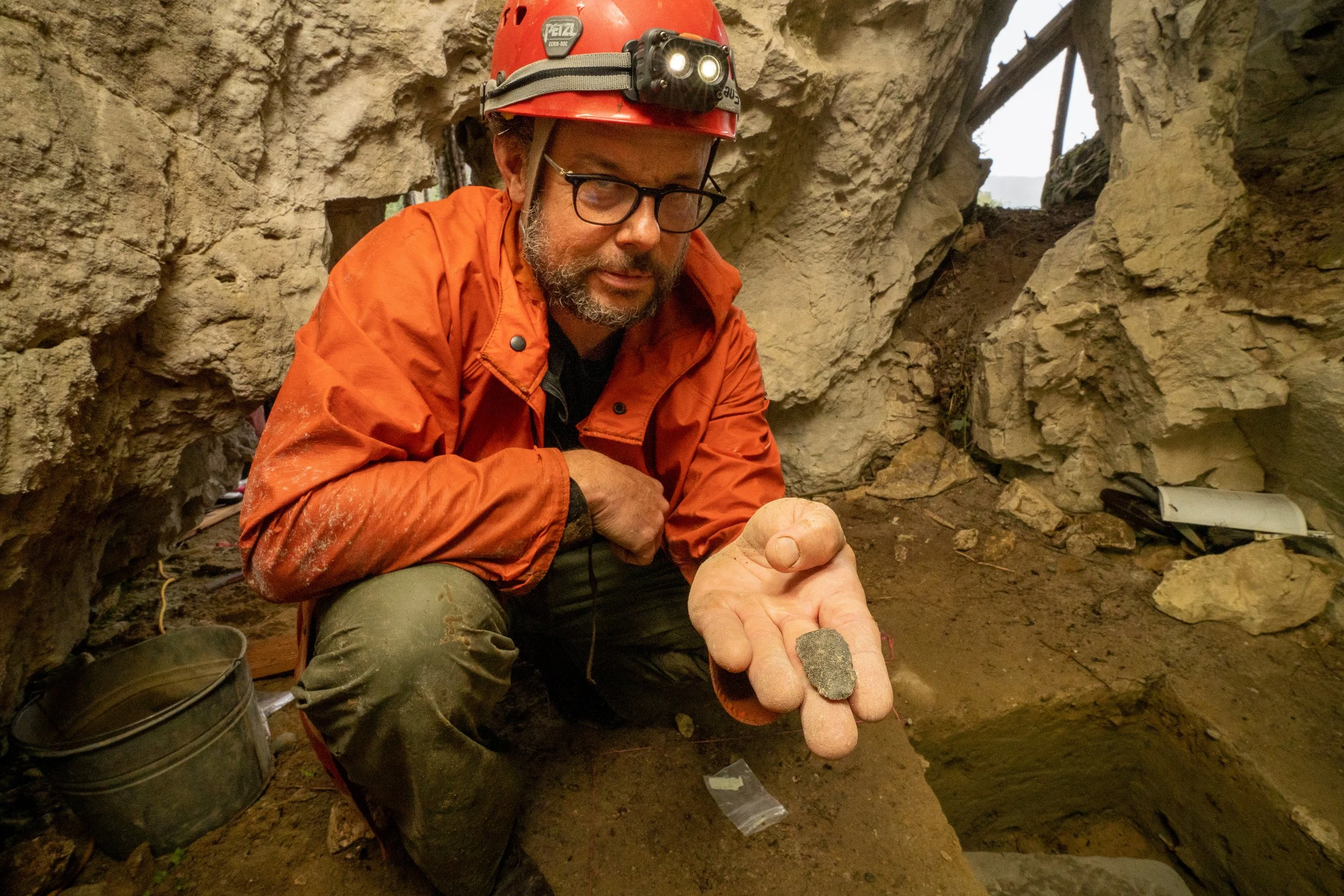 Archaeologist in orange jacket and red helmet holding a small artifact at an excavation site.