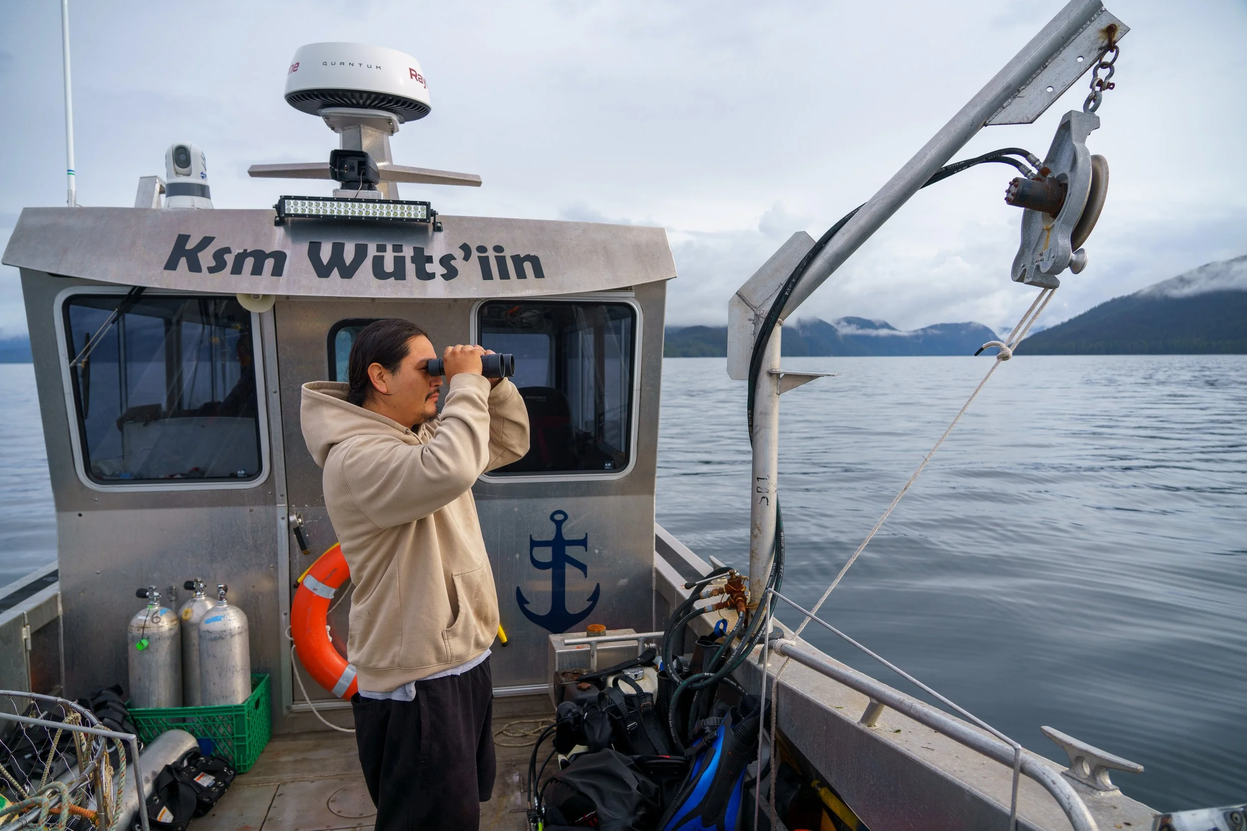 A person on a boat looking through binoculars, with a scenic view of a calm body of water and mountains in the background.