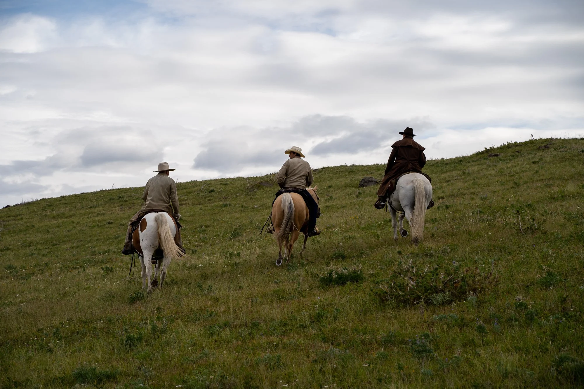 Three people riding horses up a grassy hill under a cloudy sky.