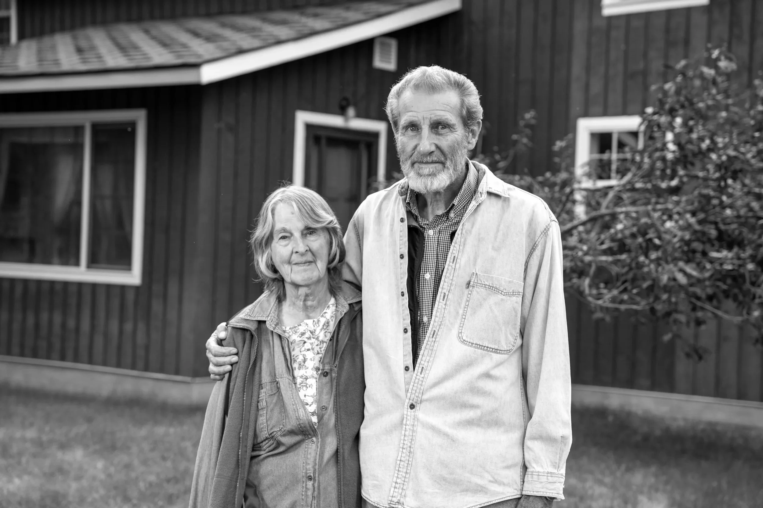An elderly couple stands outside in front of a house, with the man having his arm around the woman.