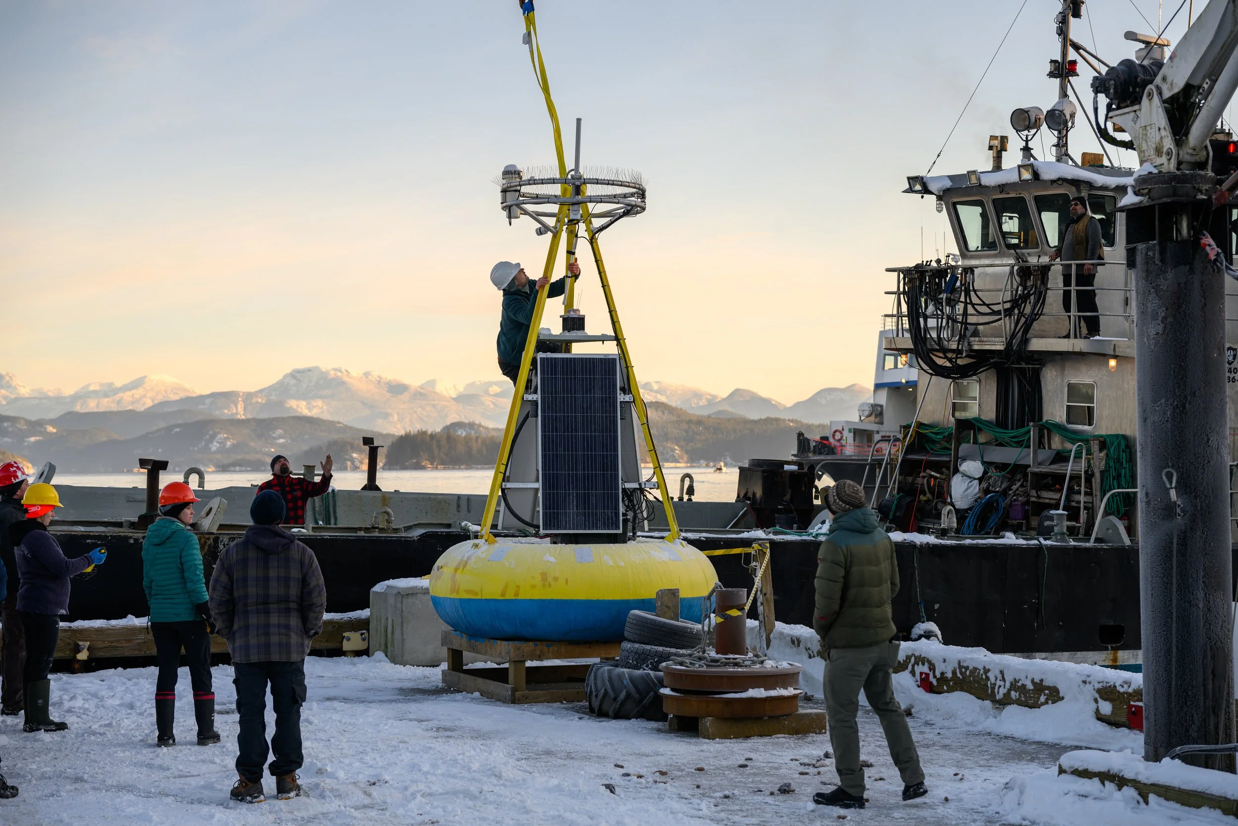 A group of people wearing winter clothing and safety helmets are on a snowy dock. One person is working on a circular device suspended from a yellow crane on a buoy, with a large ship nearby. The background features a body of water and snow-capped mo