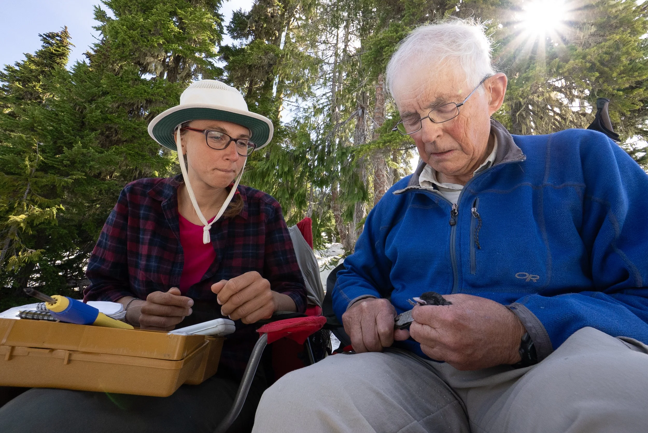 Two people, a woman and an elderly man, outdoors in a forested area, looking at tools or equipment.