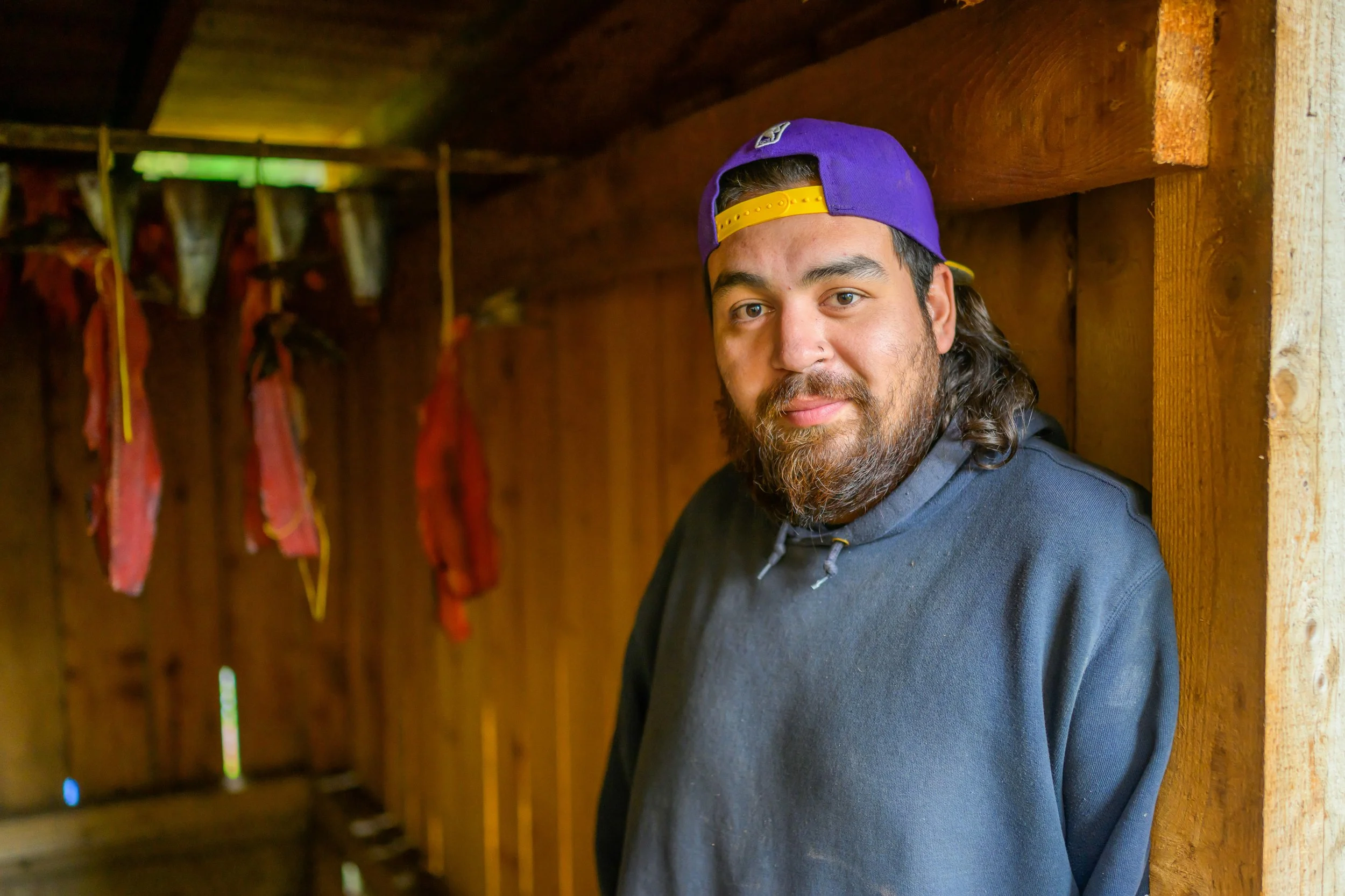 A man with a beard wearing a purple baseball cap backwards and a dark hoodie, leaning against a wooden wall inside a rustic cabin with hanging cured meats