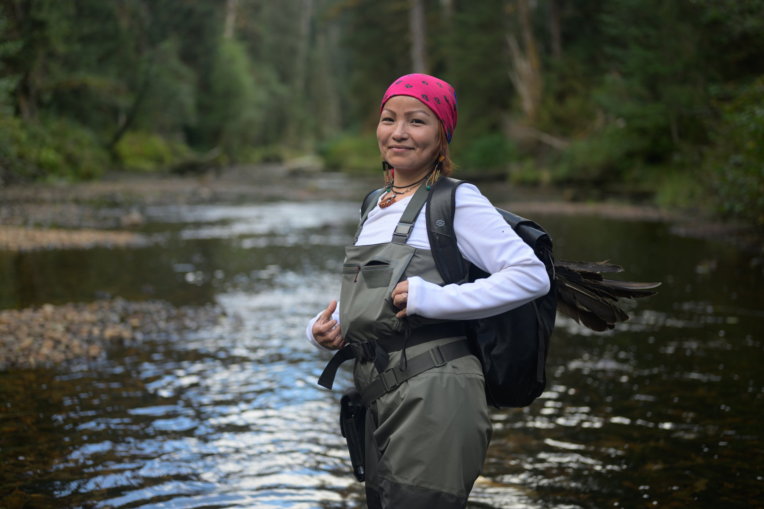 A woman standing in a river with a backpack, wearing a red bandana, white long-sleeve shirt, and outdoor gear, surrounded by trees in a forest.