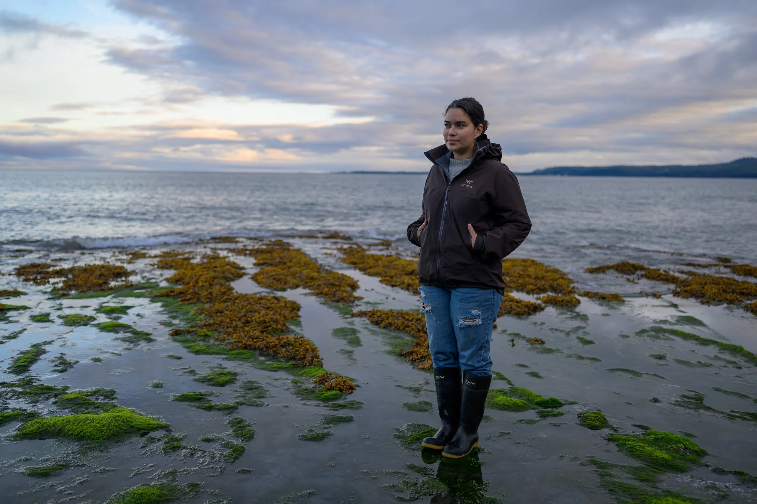 A woman standing on the rocky shoreline with green moss and seaweed, overlooking the ocean at sunset, wearing a dark jacket, ripped jeans, and black rain boots.