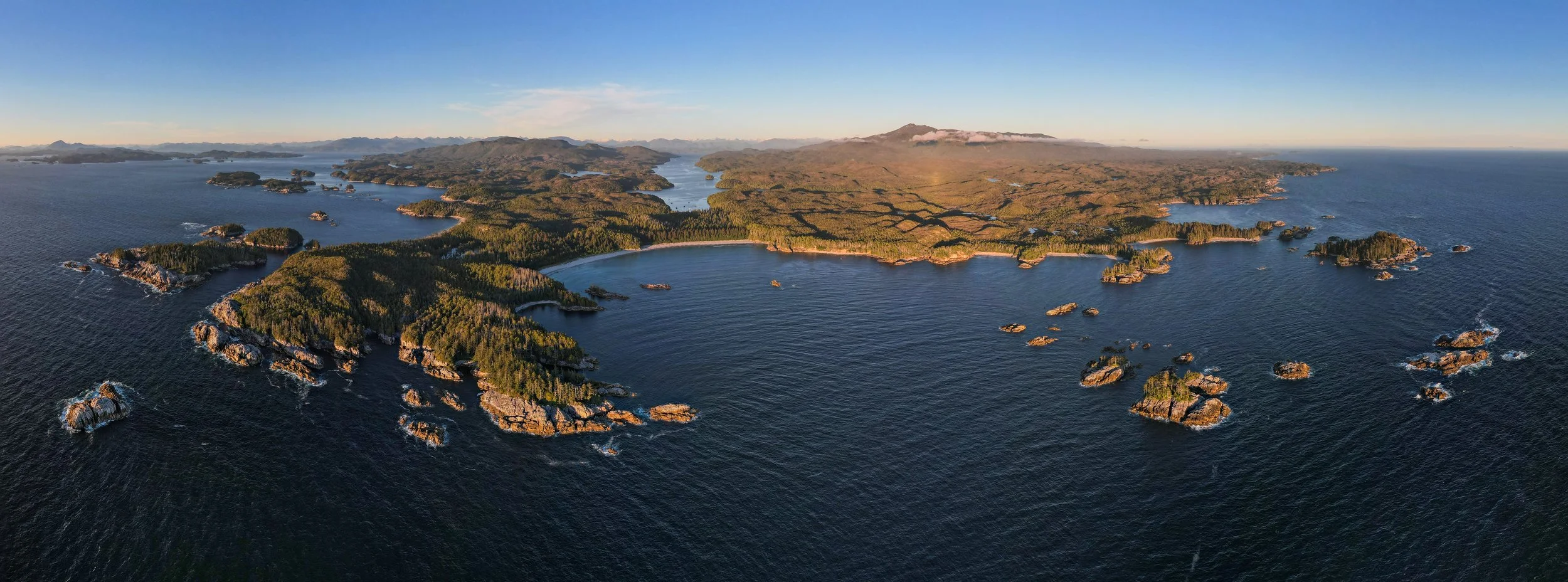 Aerial view of a rugged coastline with small islands, green forests, and a large bay, under a clear blue sky.