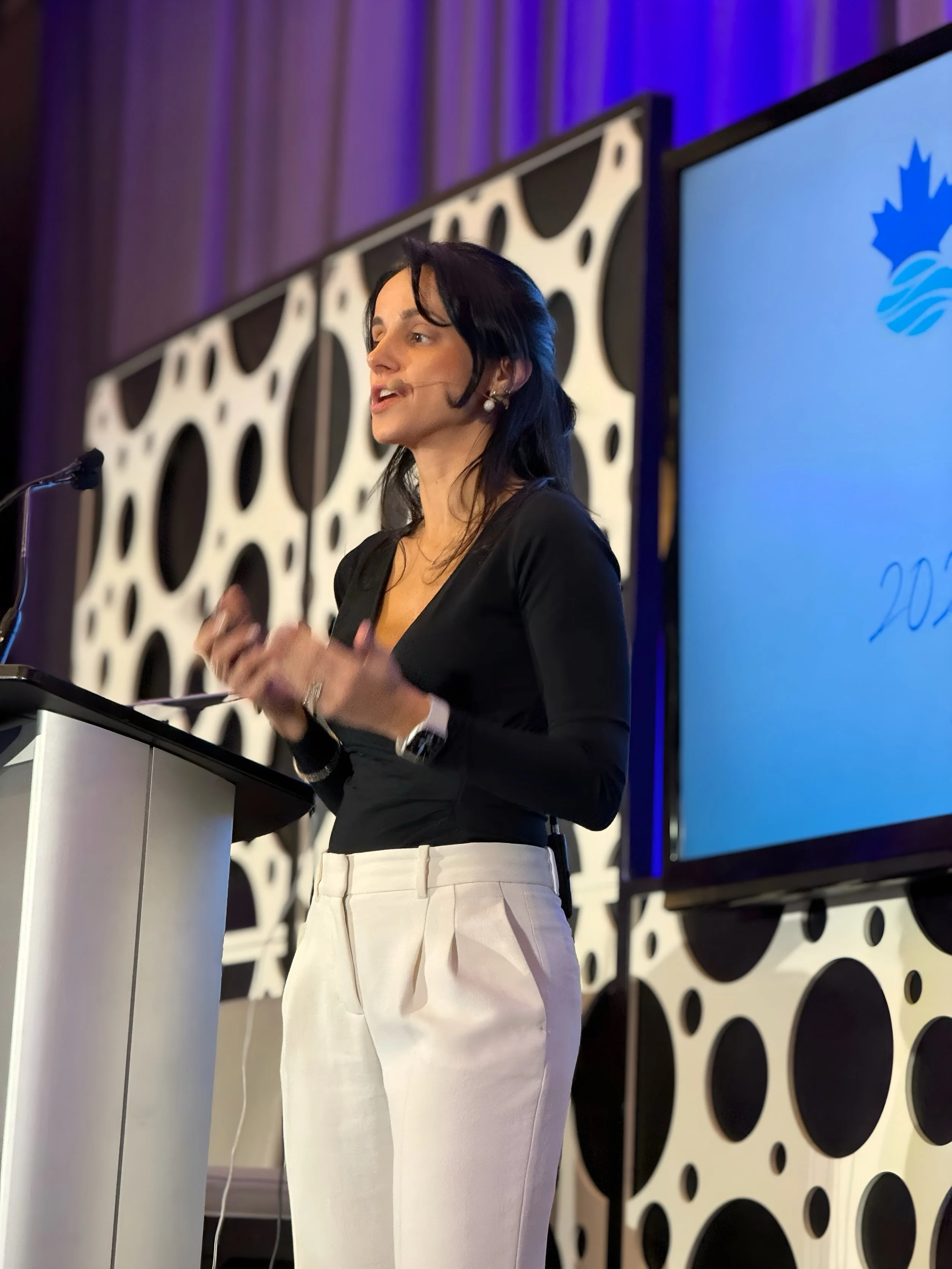 Dr. Daniela Albuquerque giving a presentation at a conference, standing behind a lectern with a microphone, with large screens displaying graphics behind her.