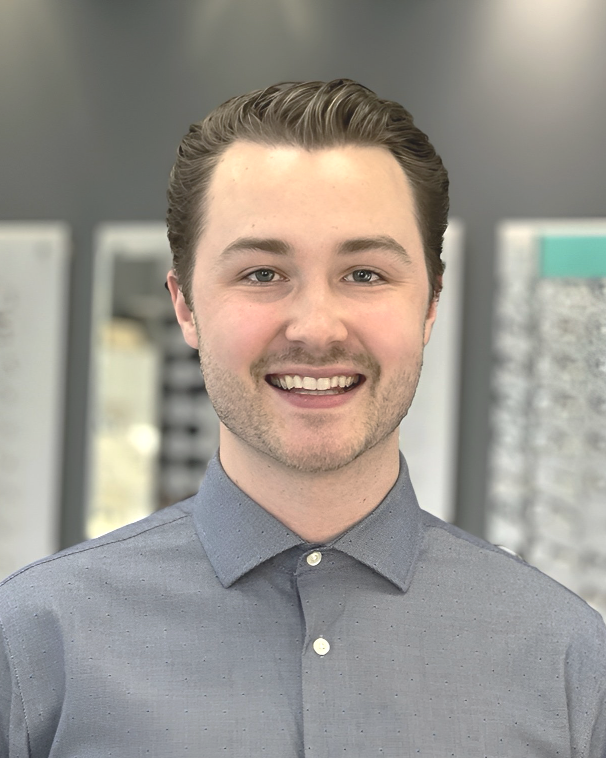 Smiling young man with light brown hair, wearing a gray button-up shirt, in an indoor setting with blurred background.