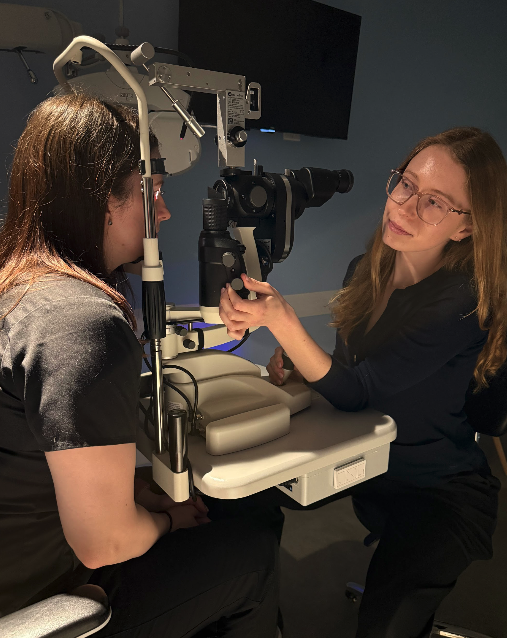 Optometrist conducting eye examination using a slit lamp while patient sits in examining chair.