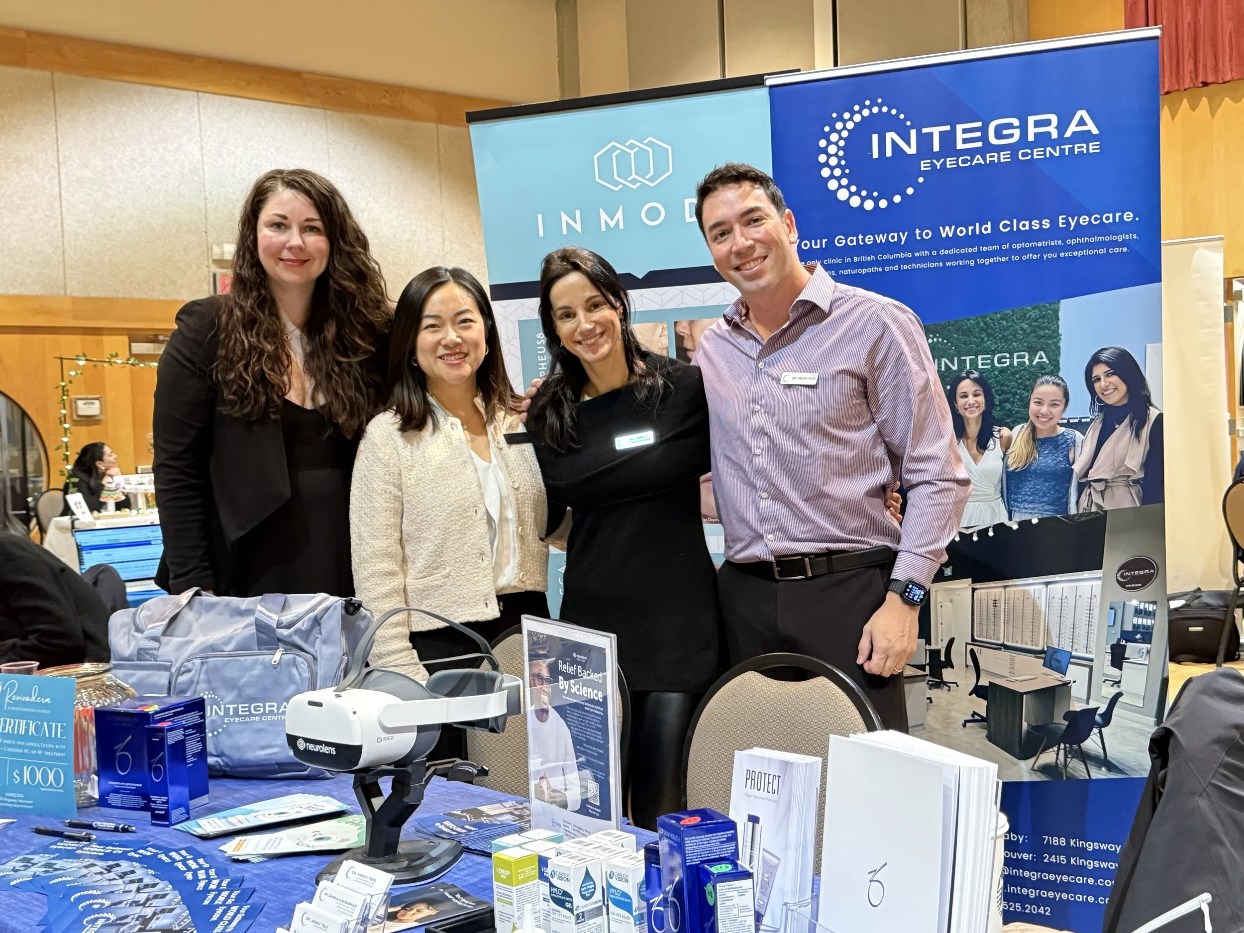 Four people standing together at a booth with a blue banner that says 'Integra Eyecare Centre' in a large room. The booth has informational materials, a virtual reality headset, and other eye care products on the table.