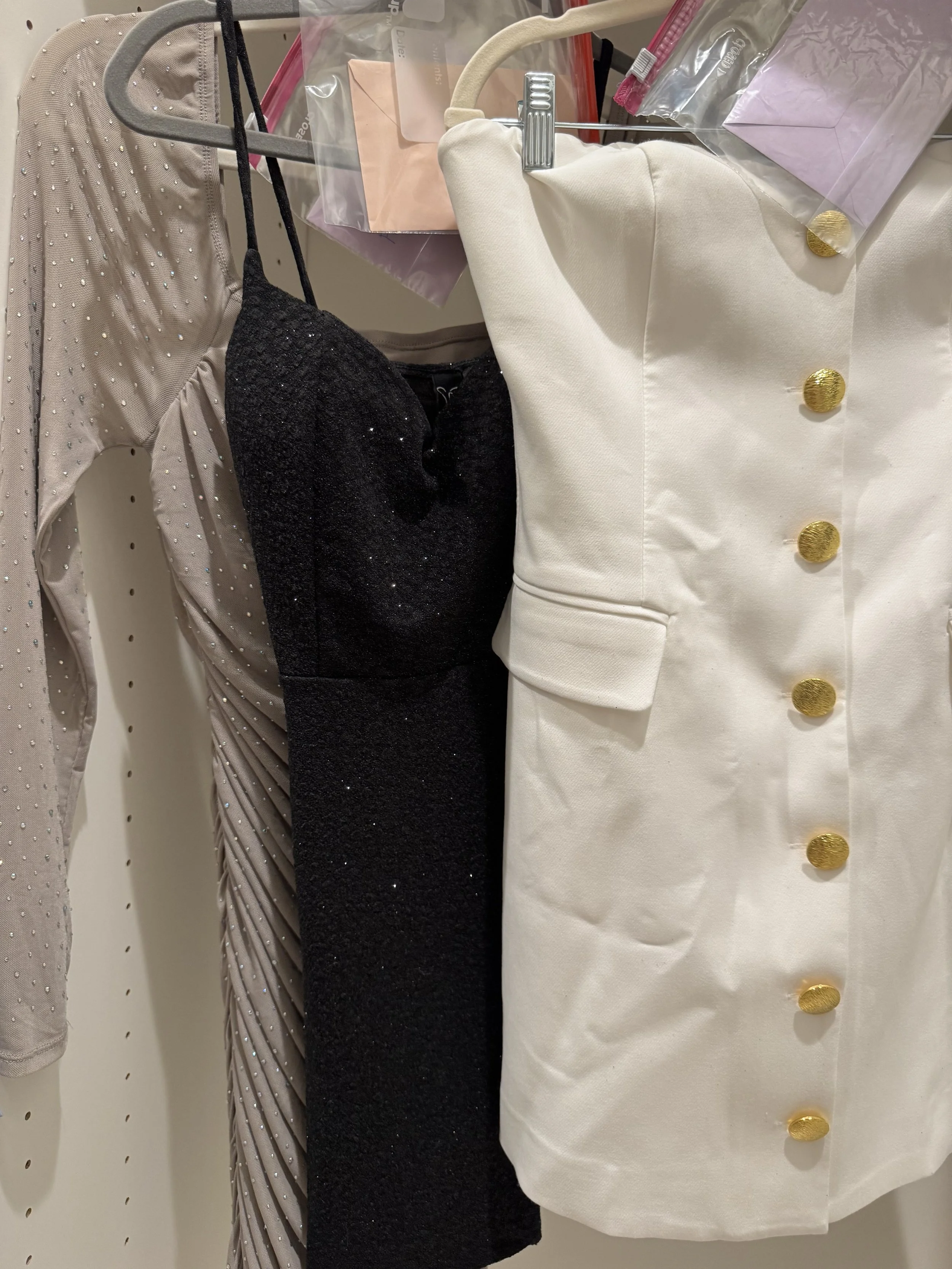 Clothing displayed on a rack, including a beige dress with rhinestones, a black dress with some sparkle, and a white coat with gold buttons.