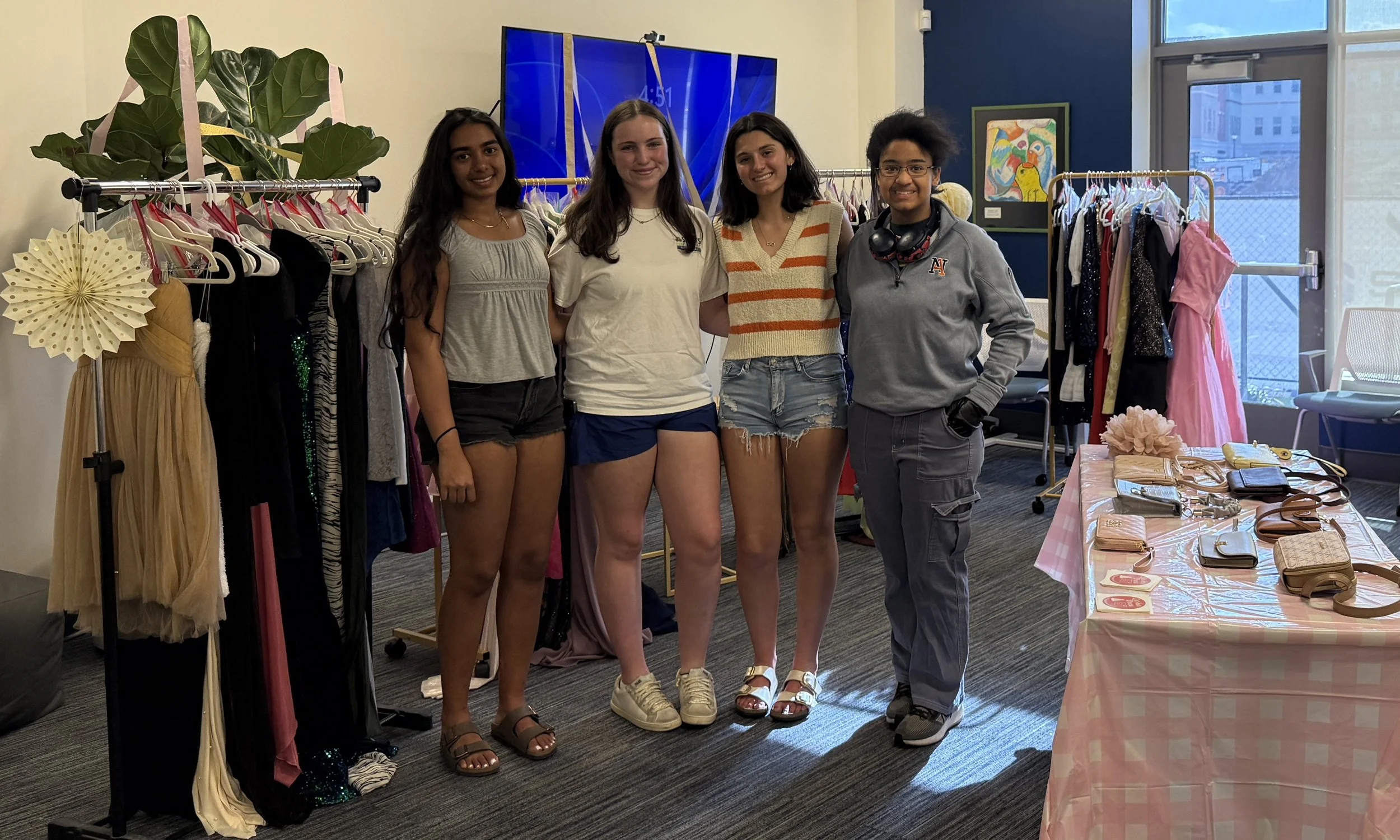 Four volunteers together inside a clothing store, smiling at the camera. Clothing racks with dresses and accessories are behind them, along with a table displaying purses and wallets. Large window letting in natural light is visible on the right.