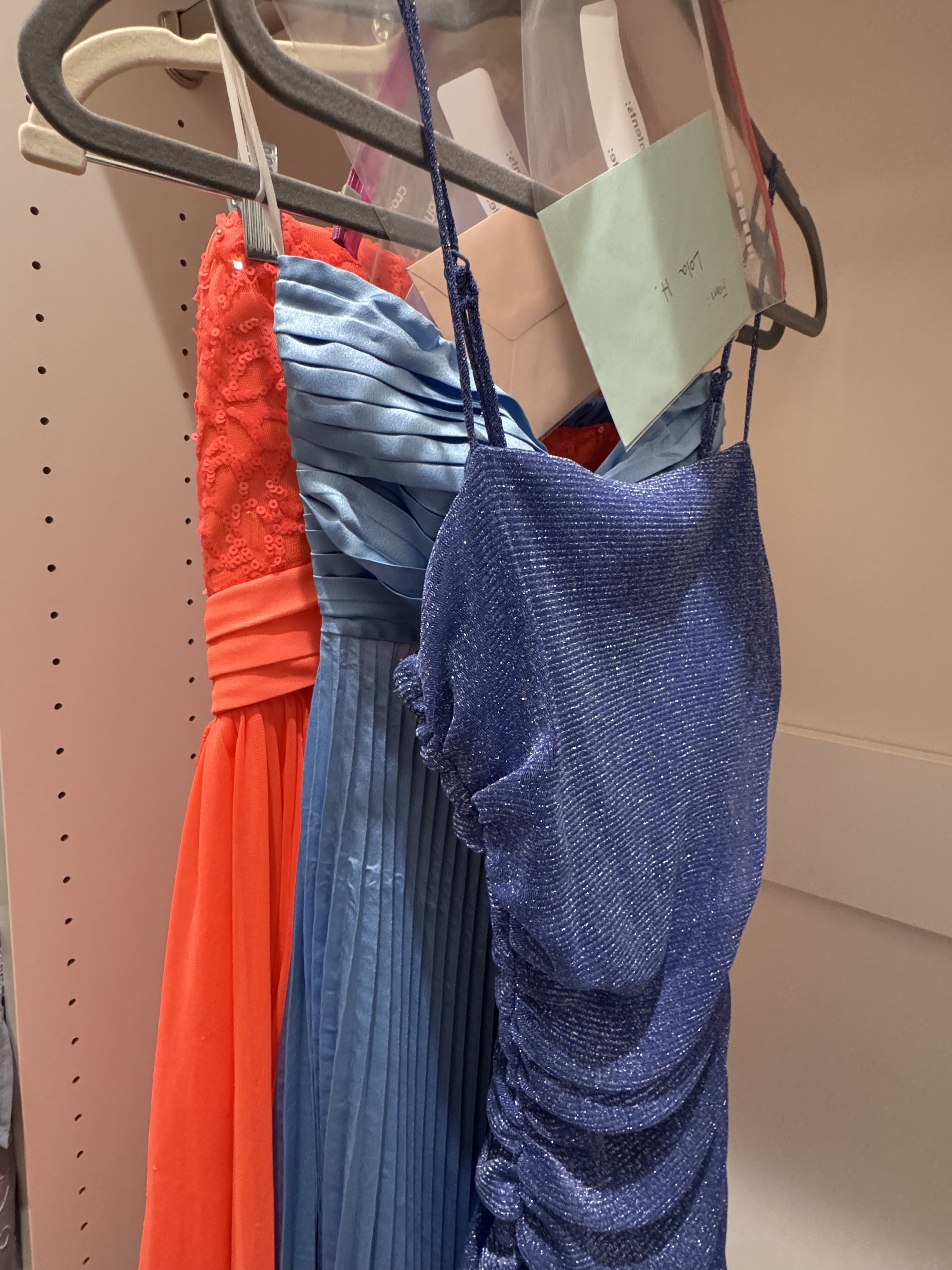 Three dresses hanging on a clothing rack. The left dress is red with sequined details, the middle dress is light blue with pleats, and the right dress is dark blue with shimmering fabric.