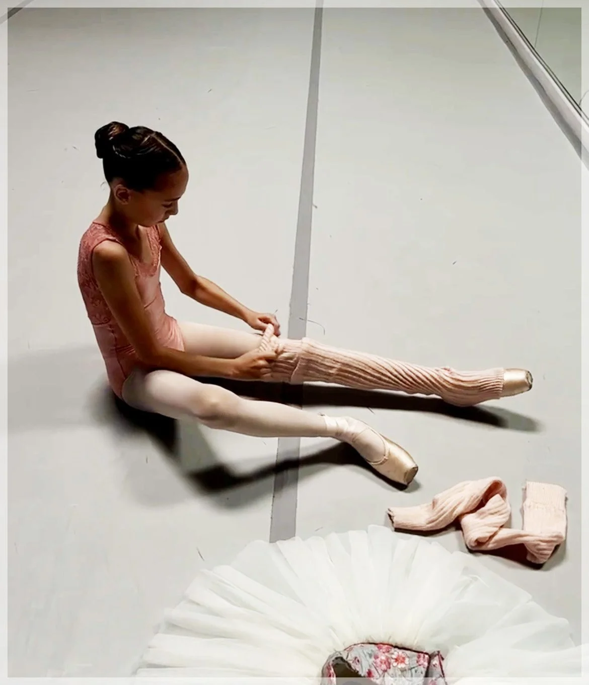 A young ballet dancer sitting on the floor putting on pink ballet tights, with ballet tutu and slippers nearby.