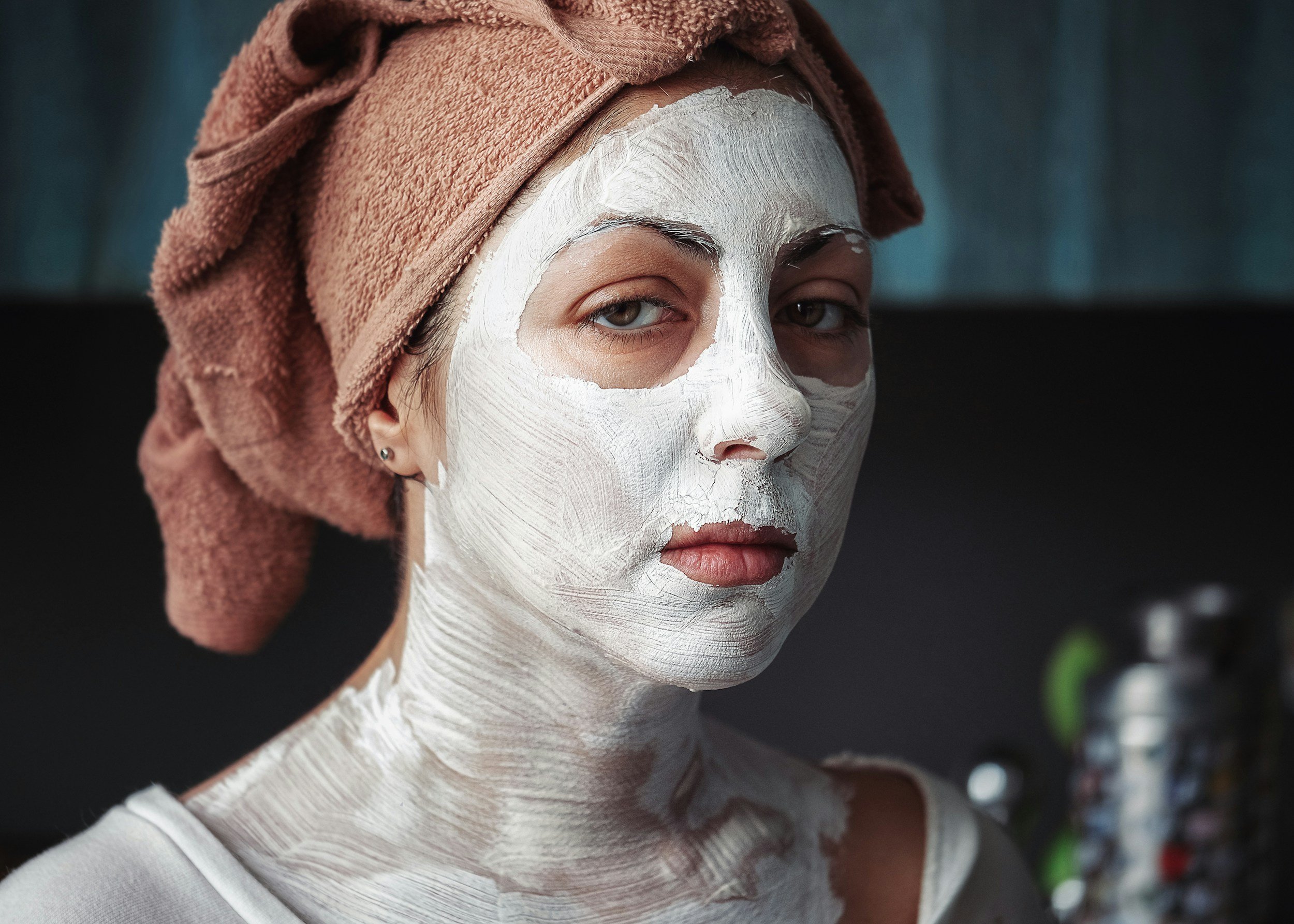 Woman with a towel wrapped around her head and a facial mask applied, looking at the camera in a bathroom or spa setting.