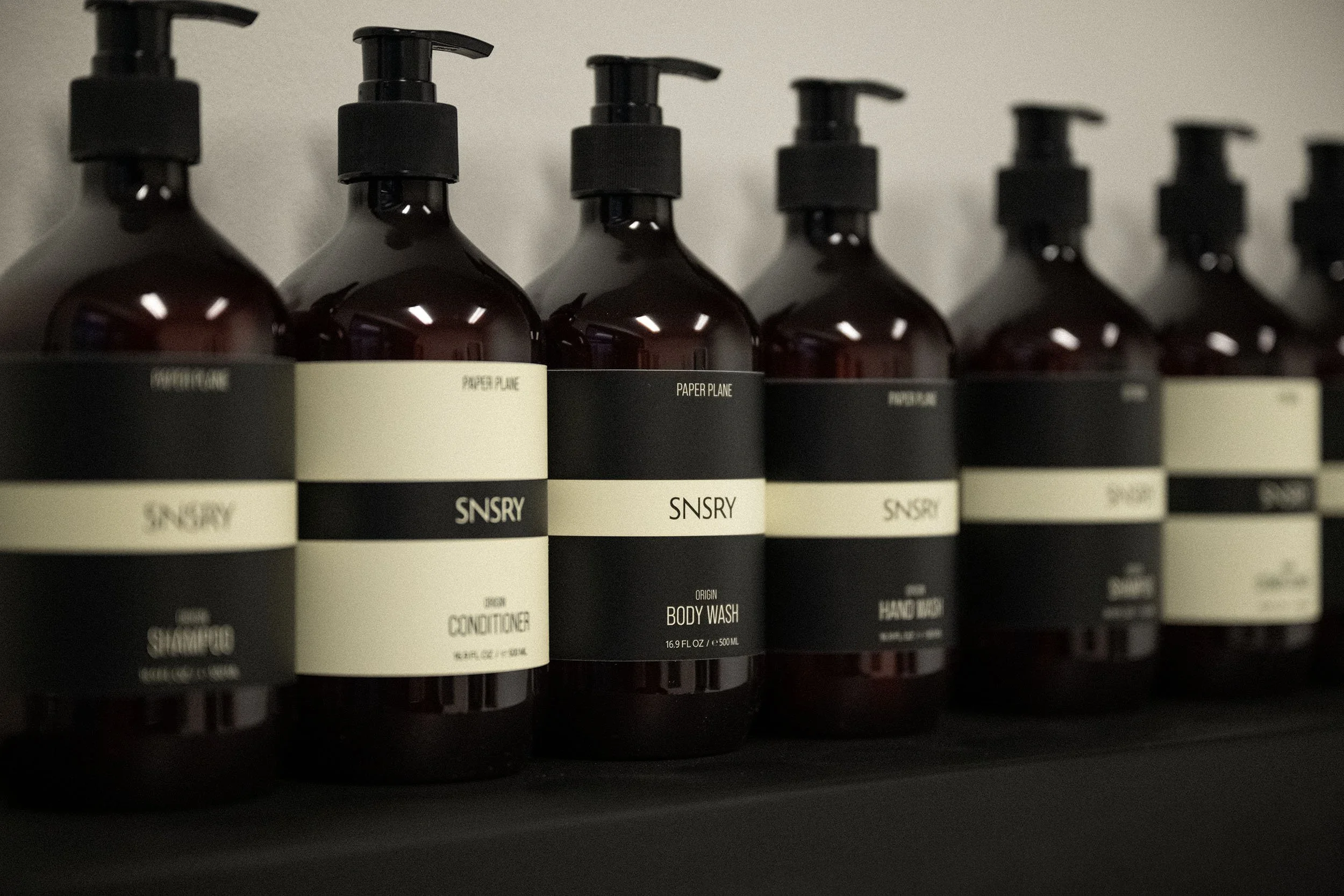 A row of dark amber bottles with black pump dispensers, labeled with minimalist black and cream labels for skincare and body wash products.