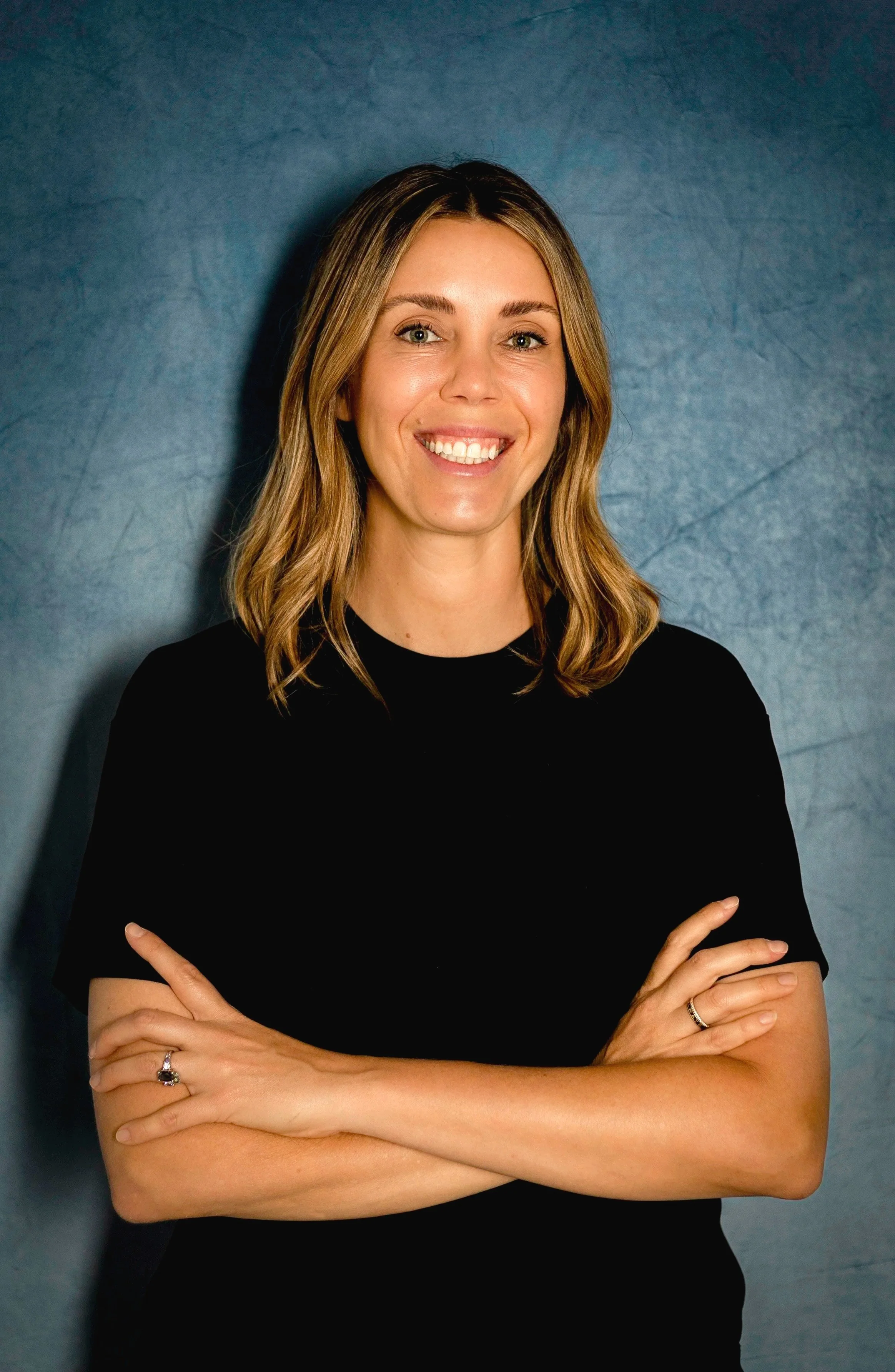 A smiling woman with shoulder-length brown hair wearing a light gray T-shirt, standing against a dark textured background.