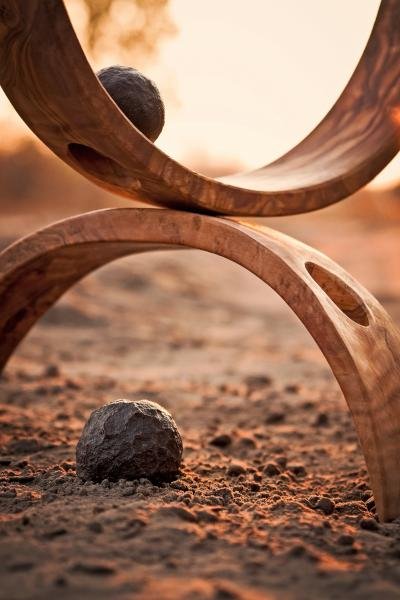 A wooden sculpture with curved openings, a ball balanced inside the upper opening, and a rock on the ground below; outdoor setting at sunset or sunrise.