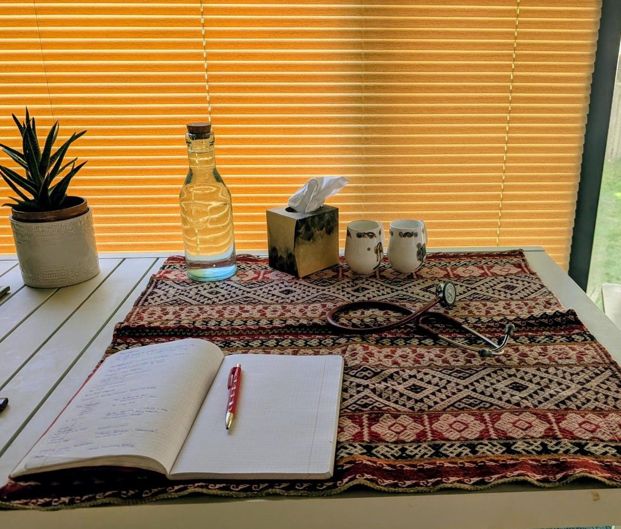 A table with a patterned cloth, a potted plant, a bottle of water, a tissue box, two ceramic cups, a stethoscope, and an open notebook with a red pen, in front of a window with closed blinds.