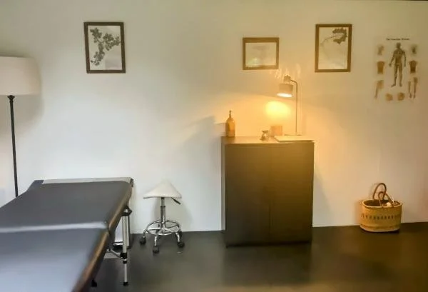 A therapy or examination room with a massage table, small stool, cabinet with lamp and decorative items, and framed artwork on a white wall.