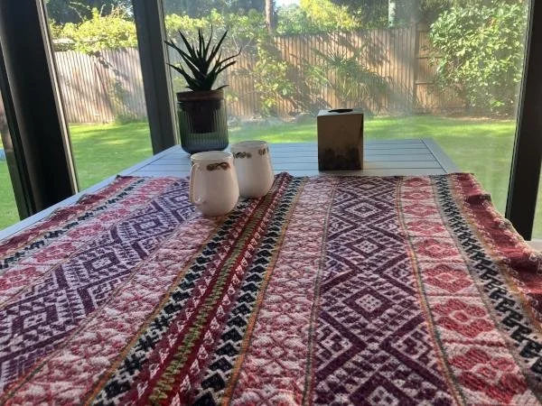 A dining table covered with a colorful, patterned cloth near large glass windows showing a backyard with grass and a wooden fence. On the table, there is a potted plant, two white candles, and a small decorative box.