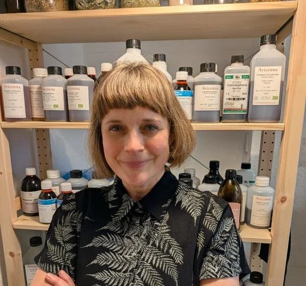 A woman with short blonde hair and a black shirt with leaf patterns, standing in front of shelves filled with bottles of medicinal or herbal products.