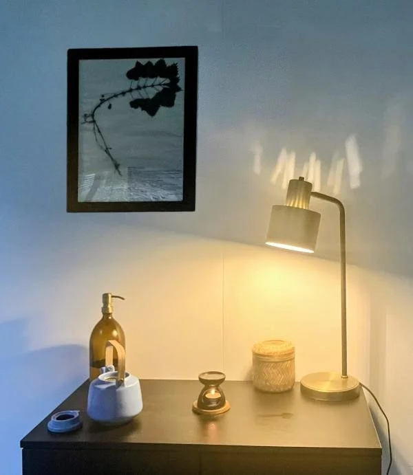 A desk with a yellowish lamp, a gold vase with a pump, a small white container, a glass hourglass, and a woven basket. A framed black-and-white botanical print hangs above the desk, with soft lighting casting shadows on the wall.