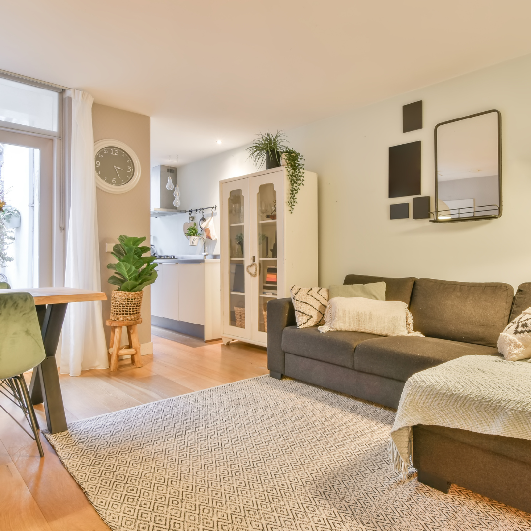 Living room with gray sofa, wall mirror, black wall art, white cabinet, dining table with green chairs, potted plant, and view into kitchen with hanging lights.