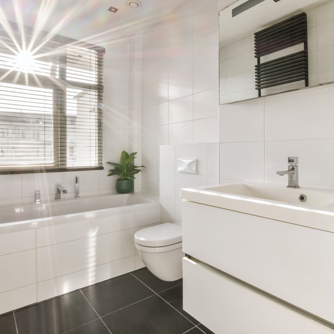 Modern bathroom with white tiled walls, a bathtub with a green potted plant, a window with blinds, a toilet, and a white vanity with a sink and mirror.