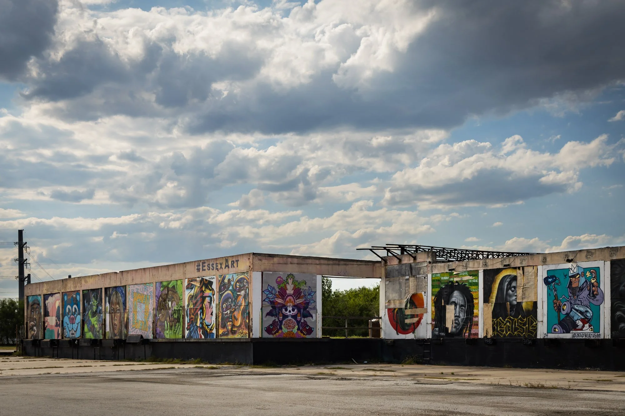 A long, partially damaged building with colorful street art murals on its side, under a cloudy sky. An empty parking lot is in foreground.
