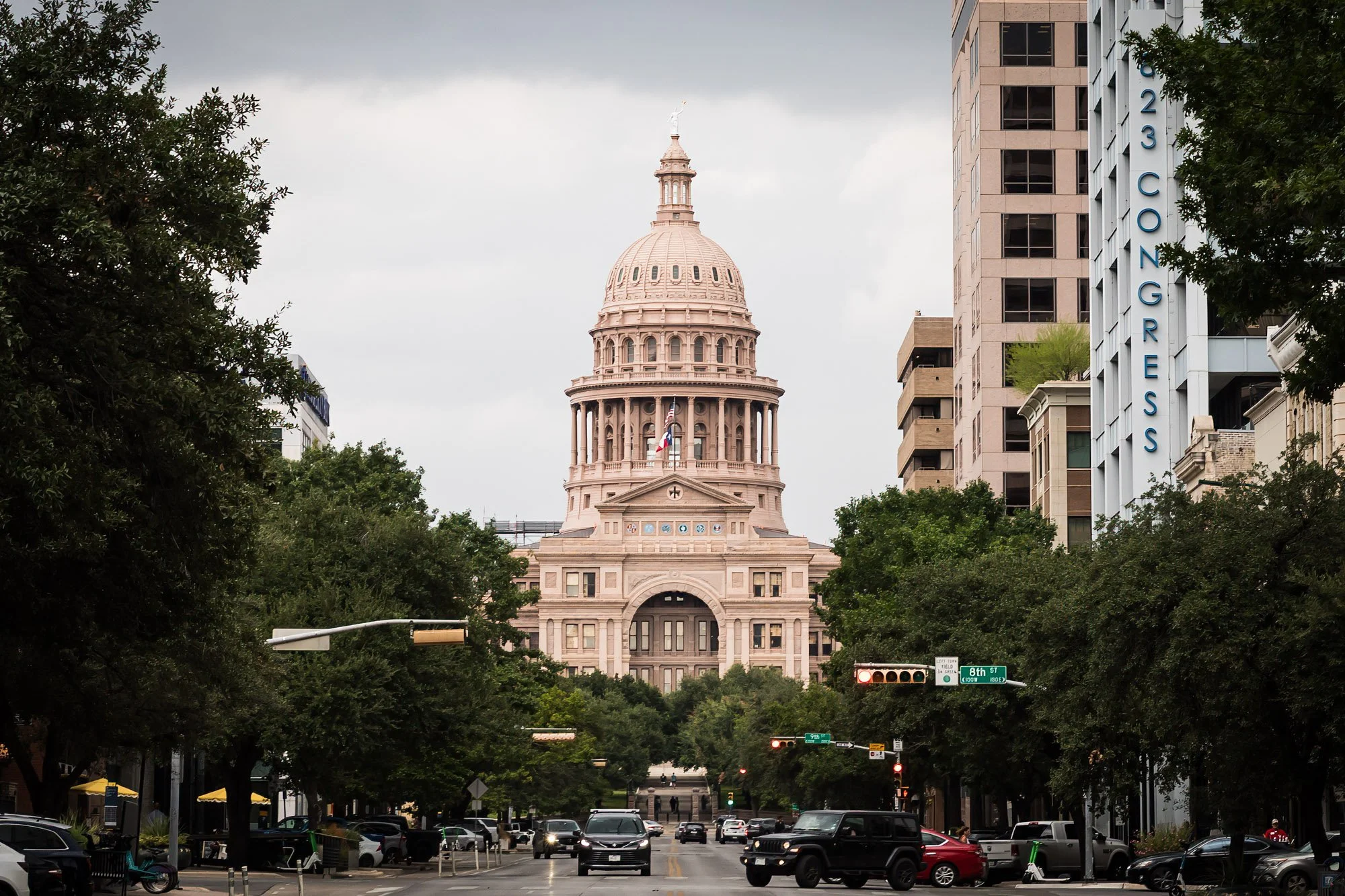 Capitol building viewed from a city street, with trees and cars, overcast sky.