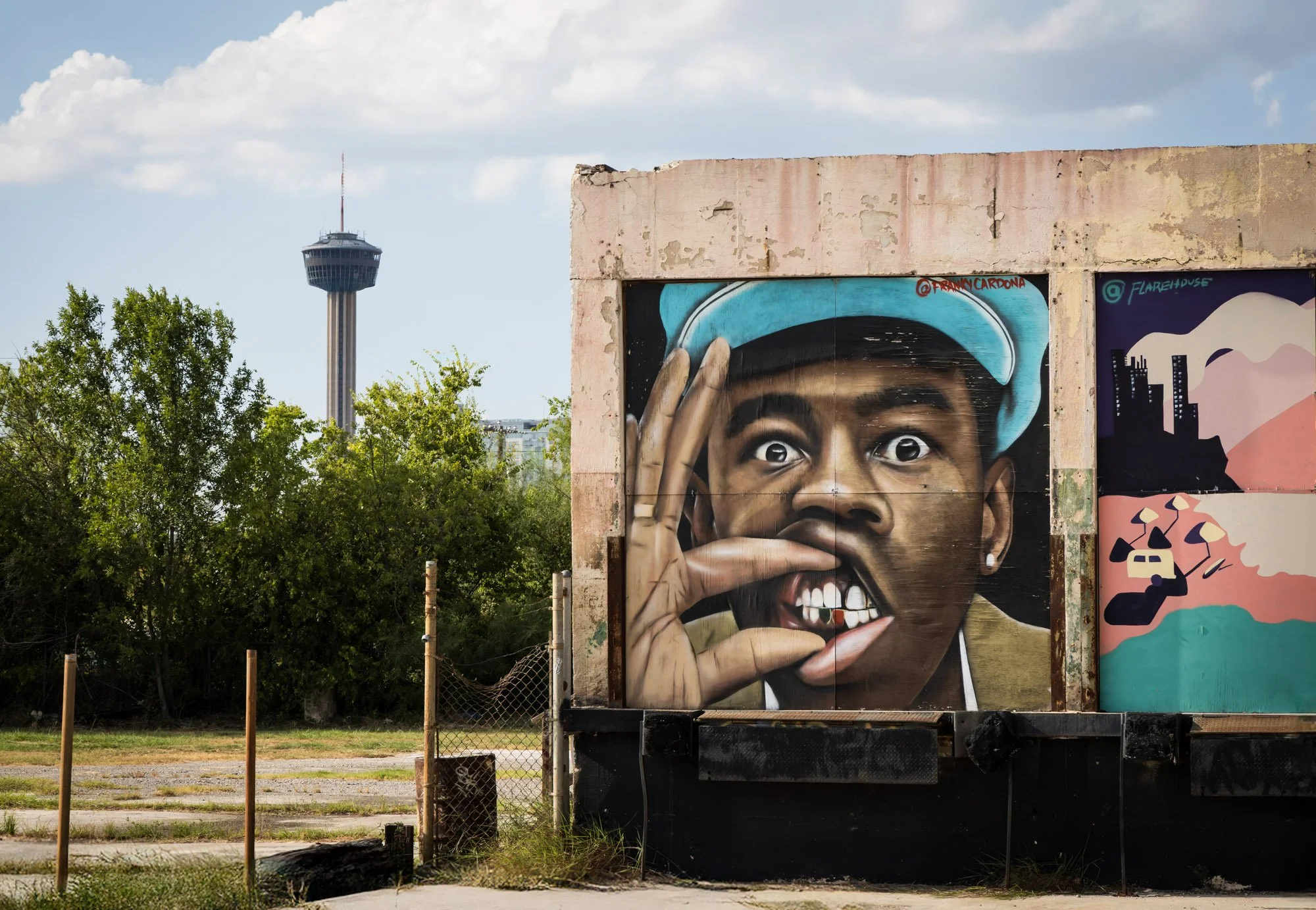 Graffiti mural of a young man with a blue cap, making an 'OK' sign near his mouth, with a surprised expression, outdoors with trees and a city tower in the background.