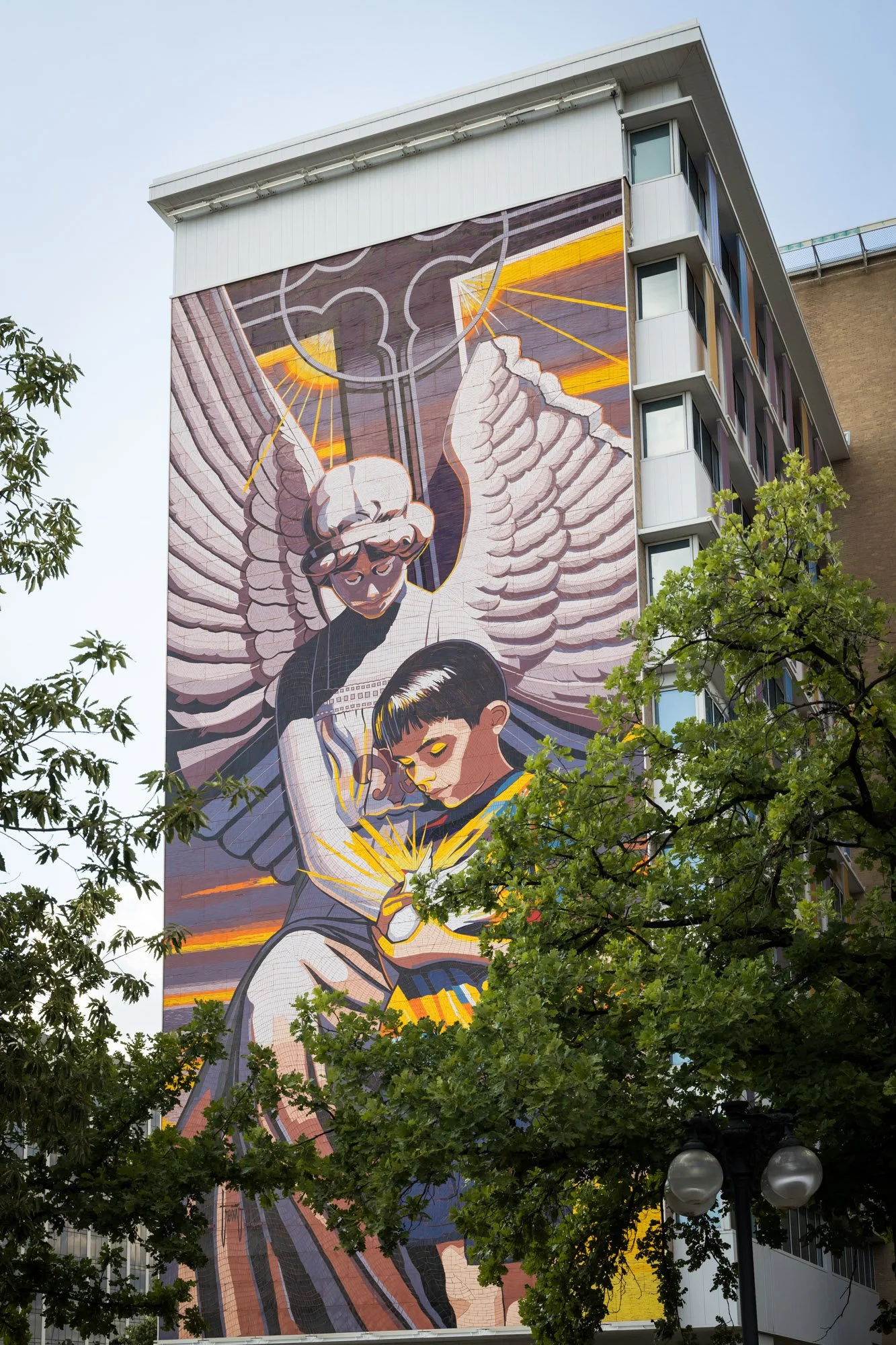 A large mural on the side of a building depicting an angelic figure with wings and a halo, descending towards a young boy with glowing eyes, holding a bright yellow object; trees partially cover the bottom of the mural.