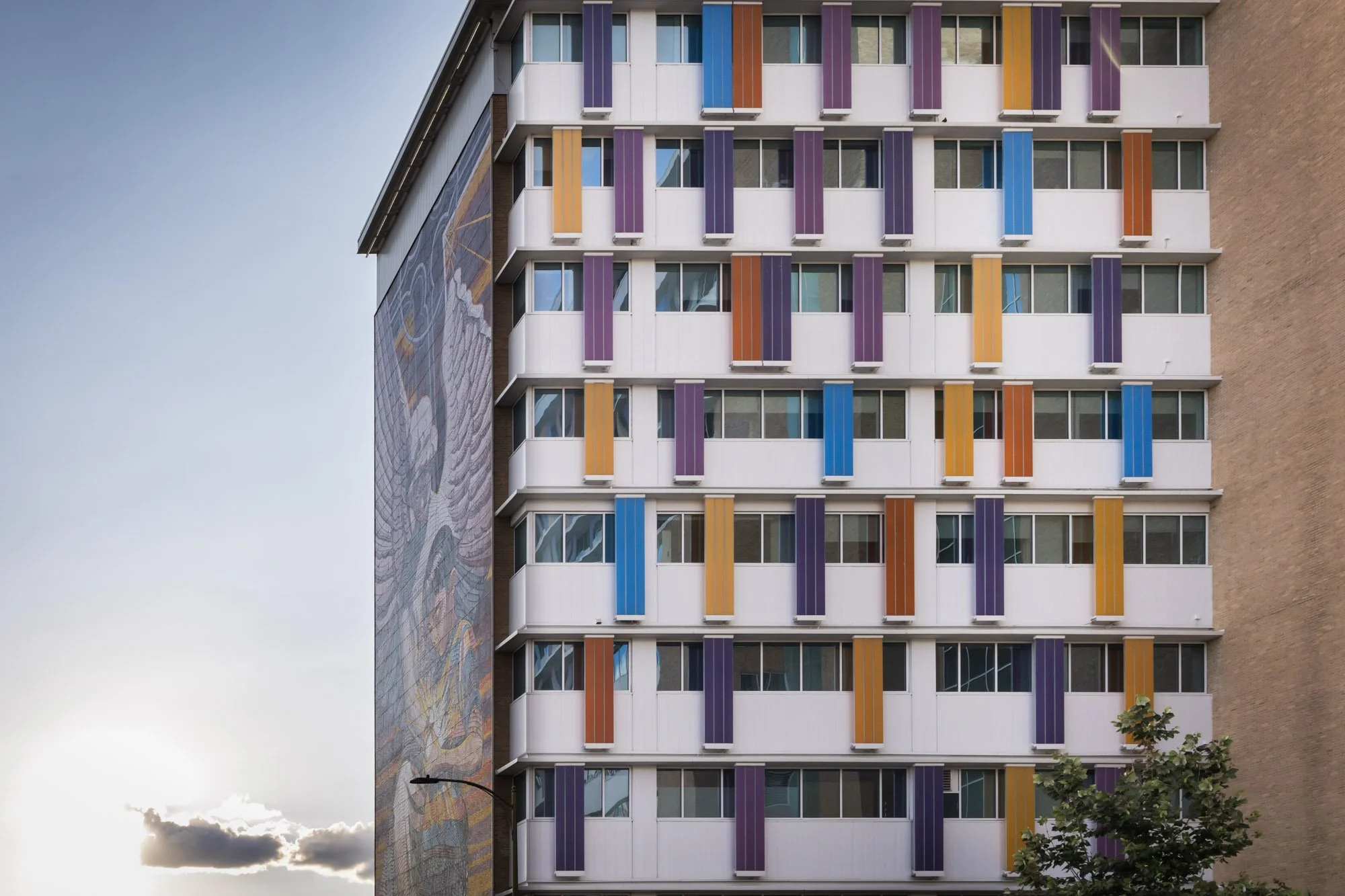 Colorful balcony panels on the exterior of a multi-story building with a mural on the side wall, a streetlamp, and a tree in the foreground, during sunset.