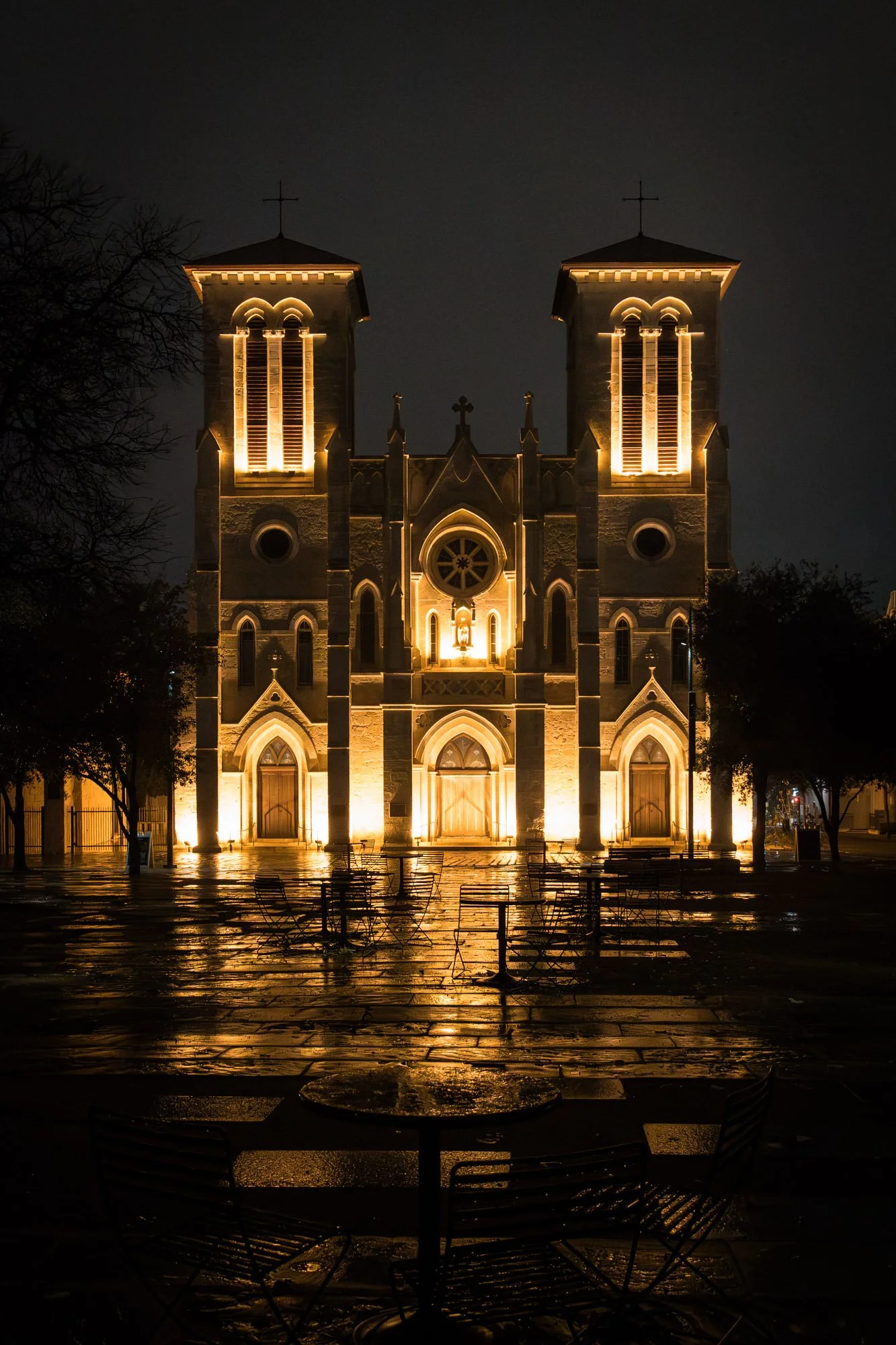 Night view of a lit-up gothic-style church with twin towers, arched windows, and altar lit behind doors, wet pavement reflecting lights, surrounded by trees and empty outdoor tables and chairs.