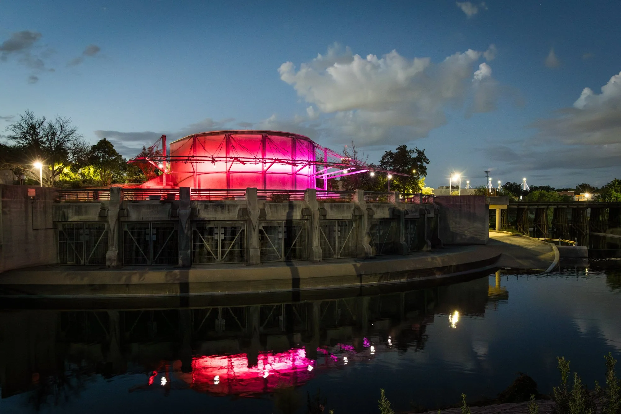 Nighttime view of a dam with a pink-lit dome structure and surrounding lights, reflecting in the water below.