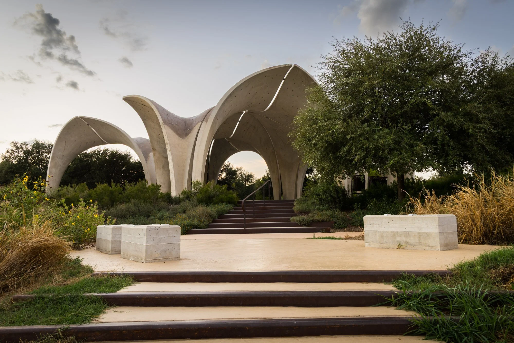 Modern concrete pavilion with curving roof supported by arching columns, surrounded by greenery and set against a partly cloudy sky.