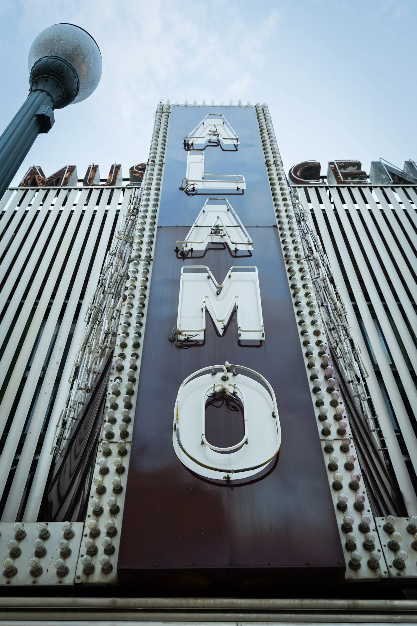 Vertical shot of a large vintage motel sign with neon letters spelling 'MOTEL' against a blue sky. The sign is framed with numerous small light bulbs, and a street lamp is visible on the left side.