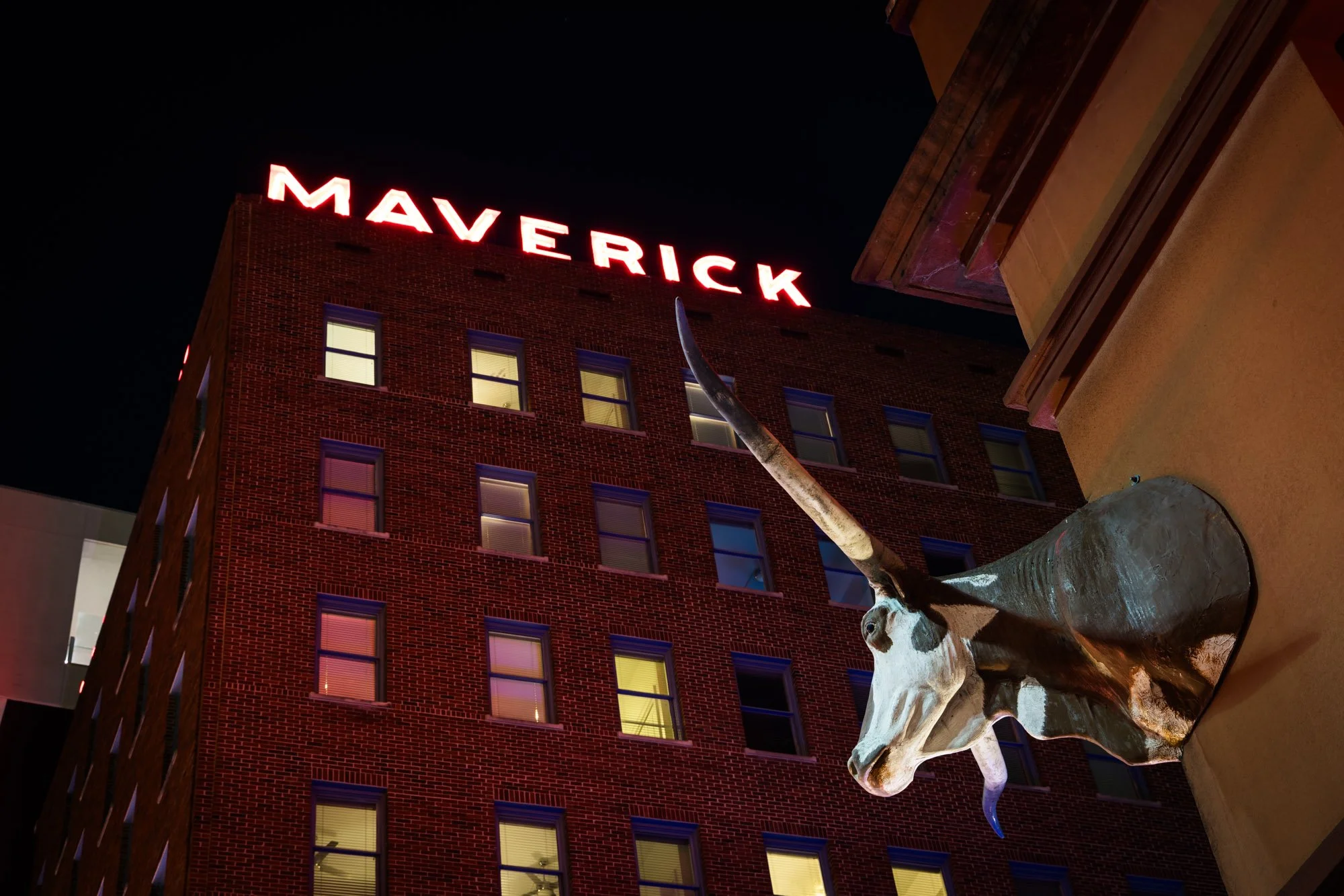 Night view of a brick building with illuminated 'MAVERICK' sign at the top. A sculpture of a unicorn's head and long horn on a wall in the foreground.