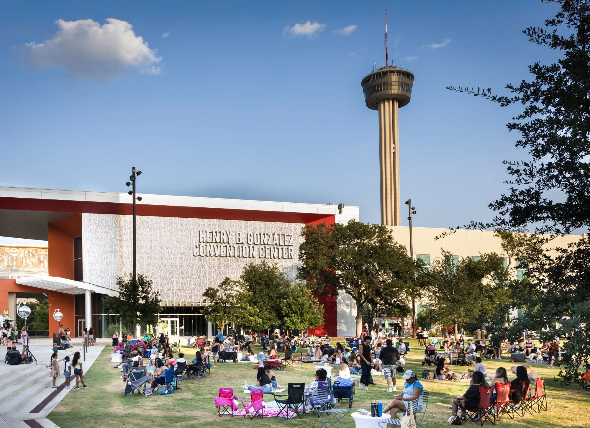 Outdoor scene at the Henry B. Gonzalez Convention Center with a crowd of people sitting on chairs and blankets on the grass area, tall observation tower in the background, trees, and a clear blue sky.