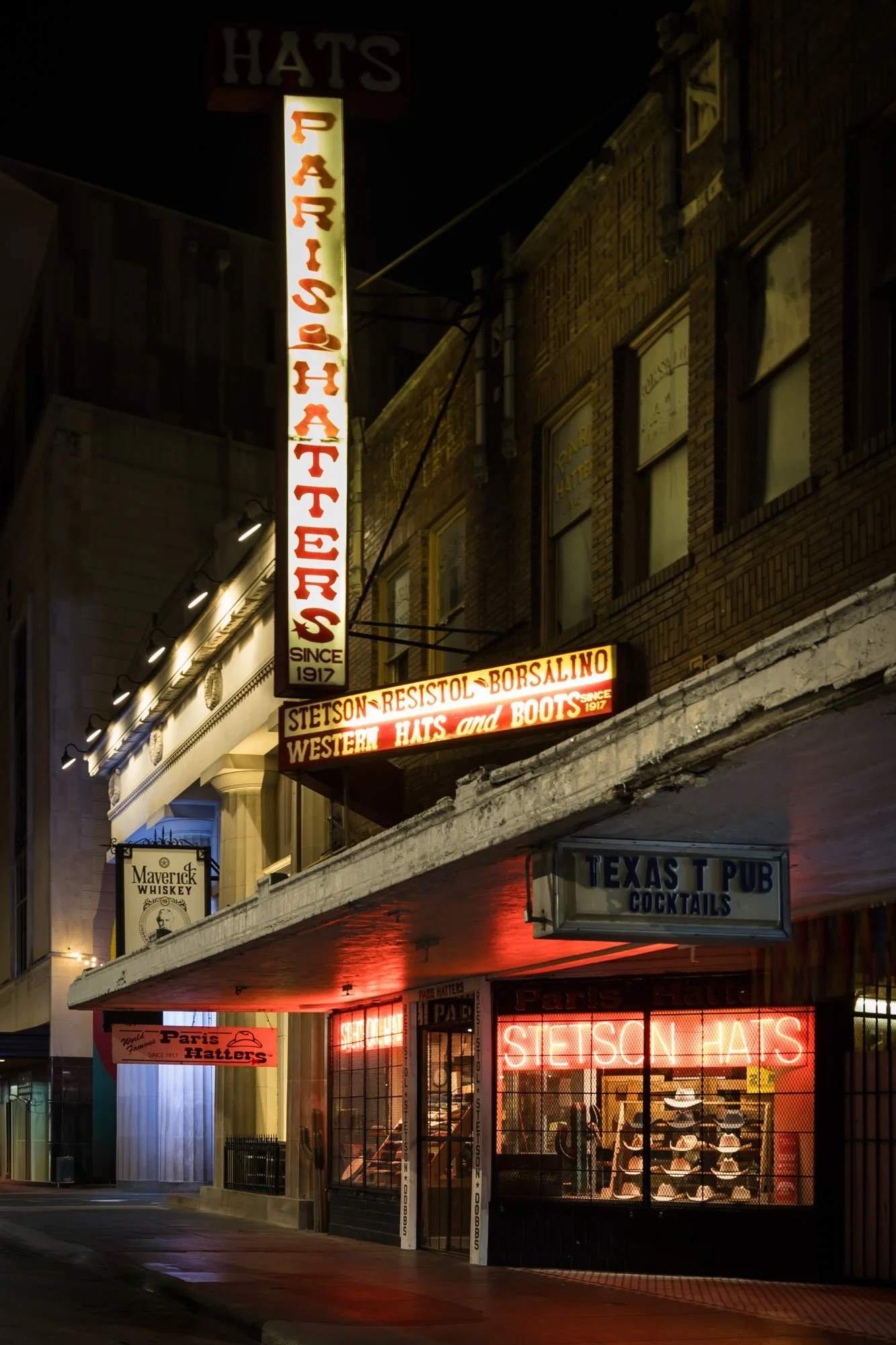 Night view of a storefront with various illuminated signs, including 'HATS', 'PARIS HATTERS SINCE 1917', 'TEXAS T PUB COCKTAILS', and 'STETSON HATS' with hats displayed inside.