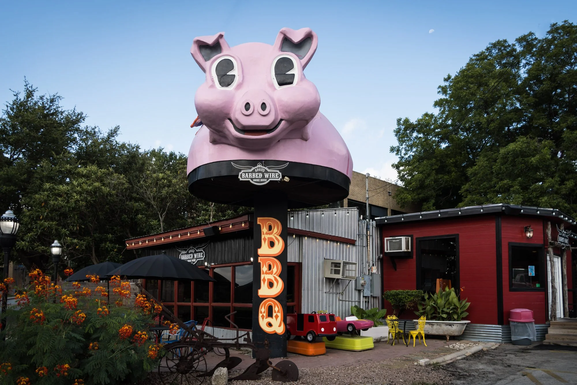 Large pink pig sculpture above a restaurant called 'Barbed Wire Smoke House', with a vertical sign displaying 'BBB' in orange letters, outdoor seating with yellow chairs, and various children's rides and toys outside.